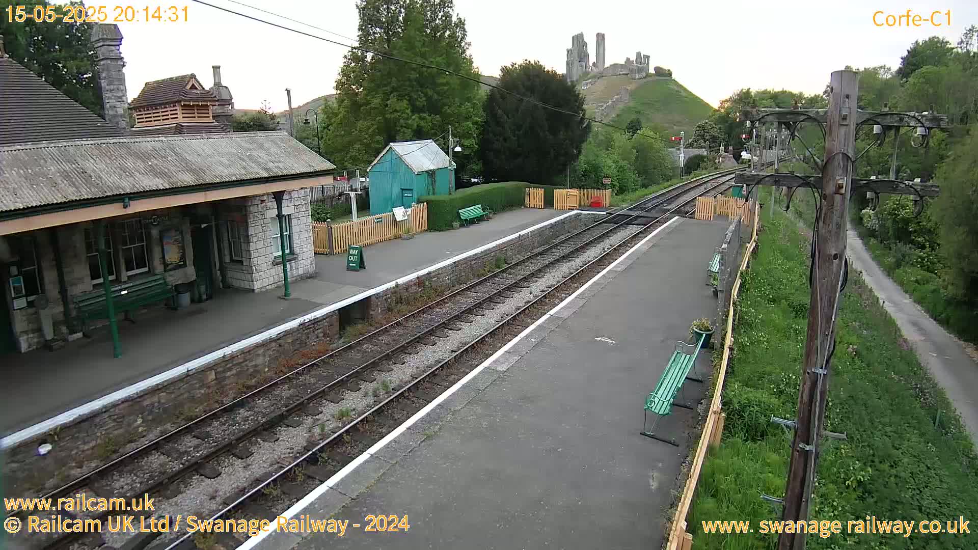A railway station platform with a stone building and a small green shed, situated near a hilltop ruin under a twilight sky.