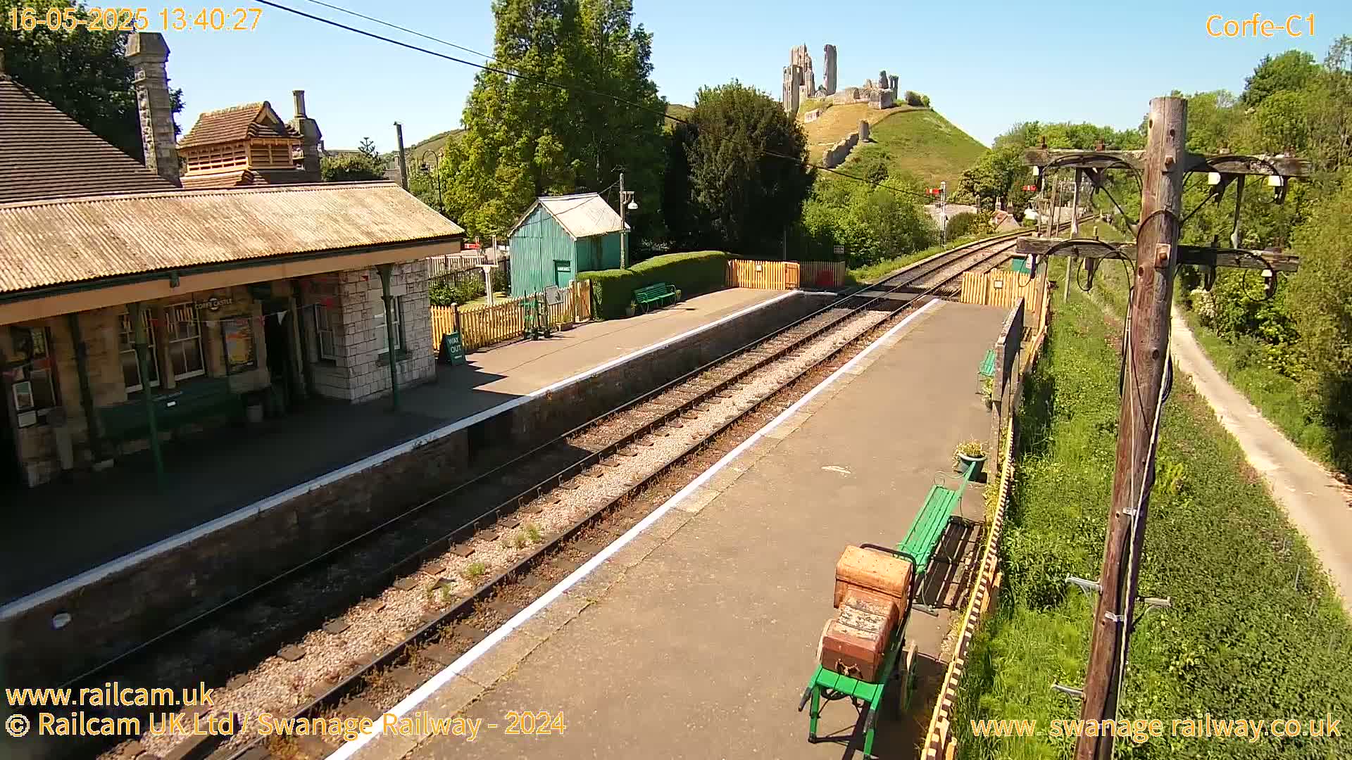 A train station platform with a stone building and a small green shed, next to train tracks, on a sunny day, with a castle ruin visible in the background on a hill.