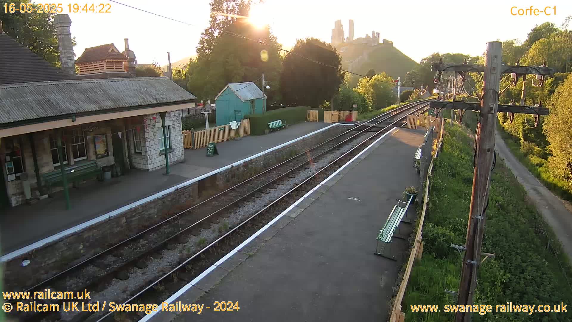 A train station platform with a stone building and a distant castle is bathed in the golden light of a setting sun.