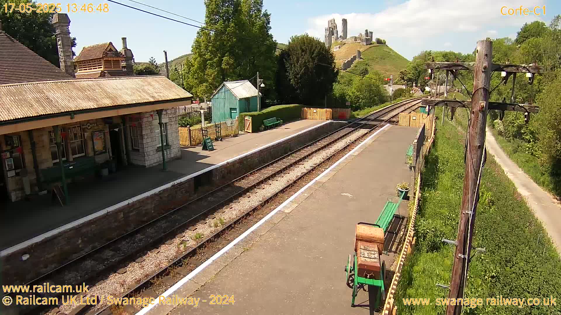 A train station platform with a stone building, a small green shed, and a distant view of a castle ruin on a hill under a sunny sky.