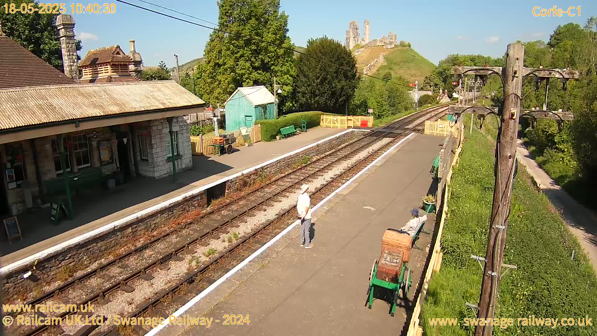 A train station platform on a sunny day, with a stone building, a small green shed, and a distant ruined castle visible on a hillside.
