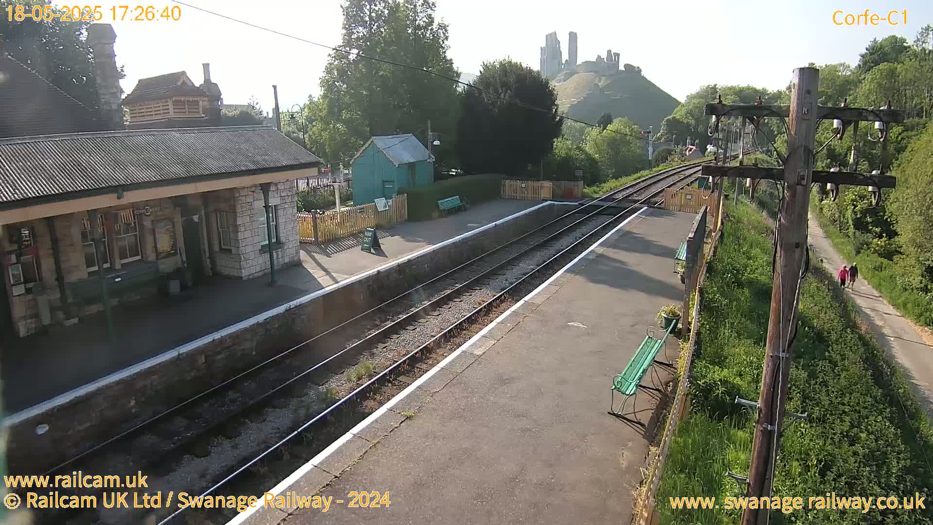 A train station platform with a stone building, a small green shed, and a distant hilltop castle is visible on a sunny day.