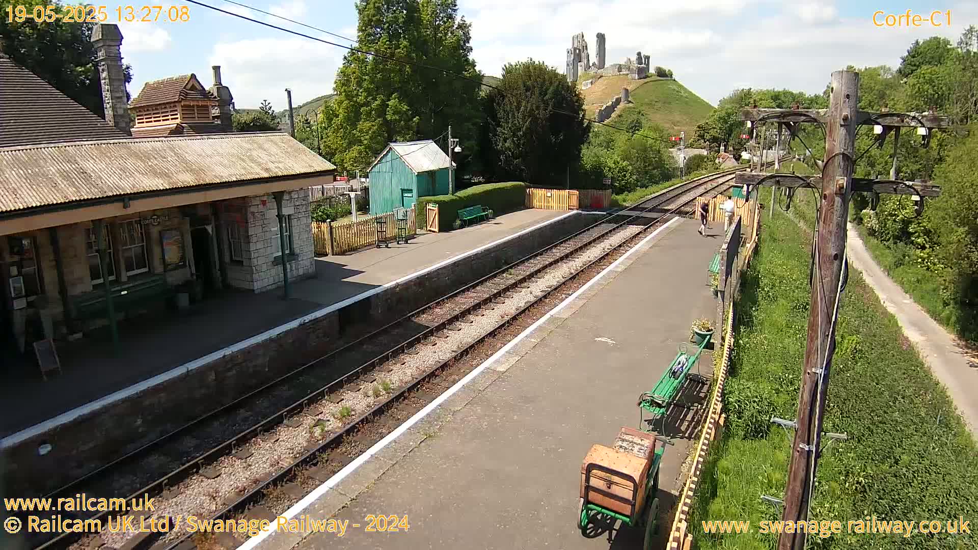 A train station with a stone building and a small green shed, situated near a hill topped with castle ruins under a partly sunny sky.