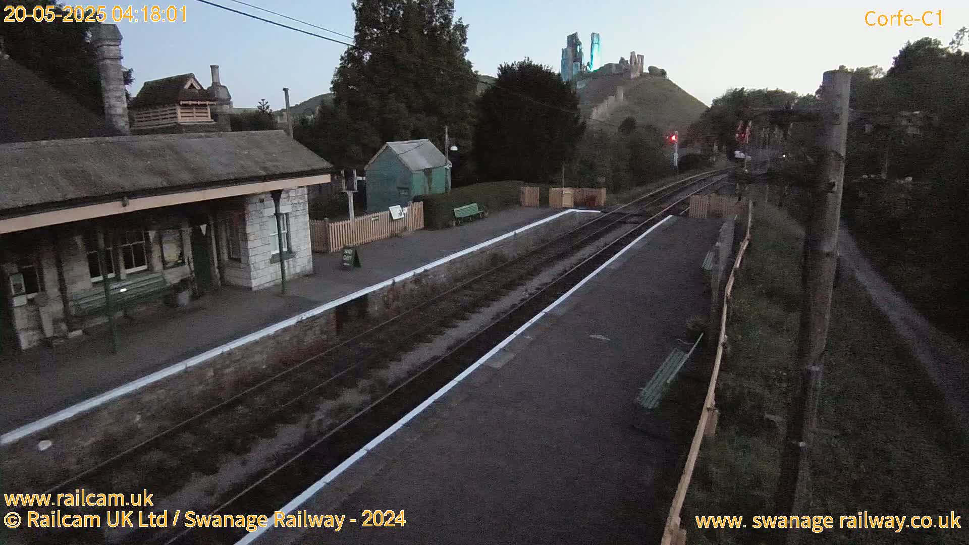 A train station platform with a stone building and a castle visible on a hill in the background under a twilight sky.