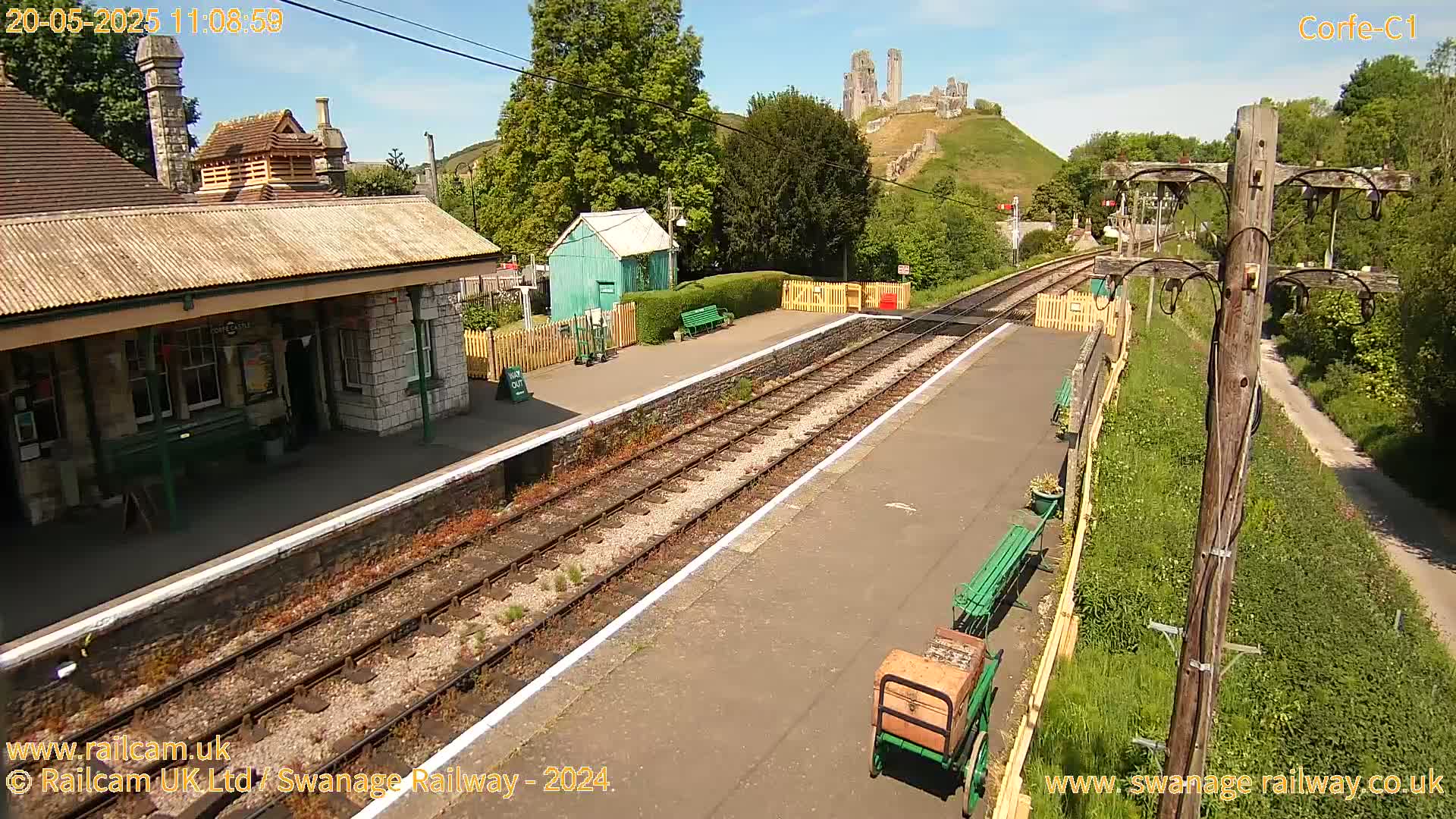 A train station with a stone building, a small green shed, and a distant castle ruin on a hill under sunny skies.