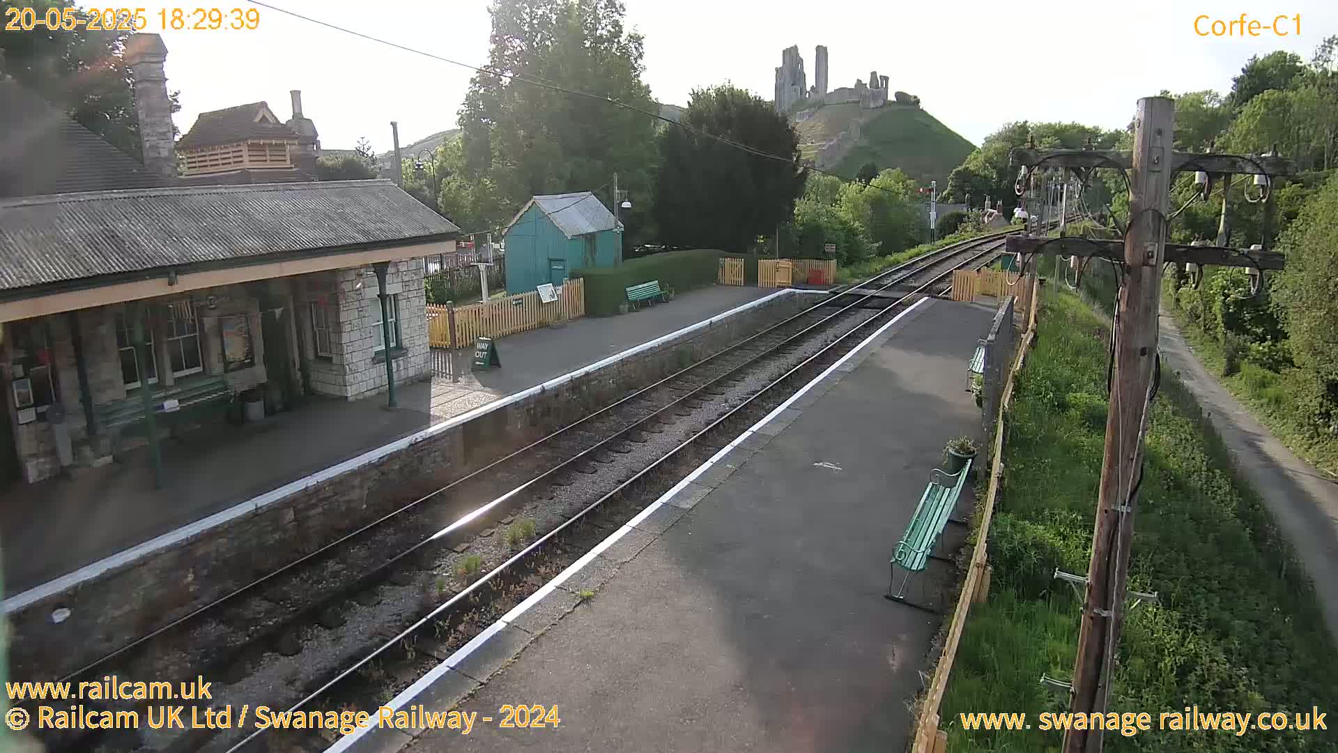 A train station platform with a stone building and a small green structure, overlooking a hilltop castle ruin under a sunny sky.