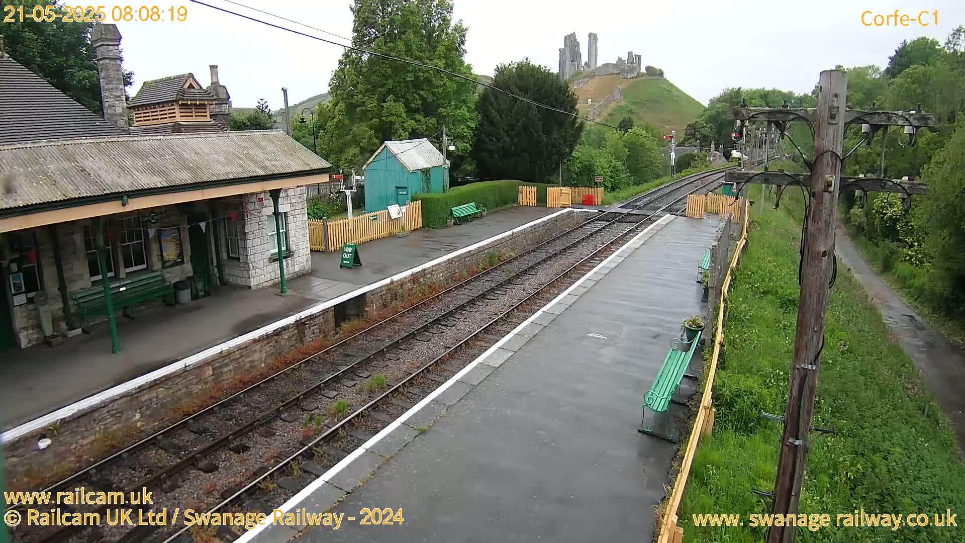 A railway station platform with a stone building, a small green shed, and a distant ruined castle visible under an overcast sky.