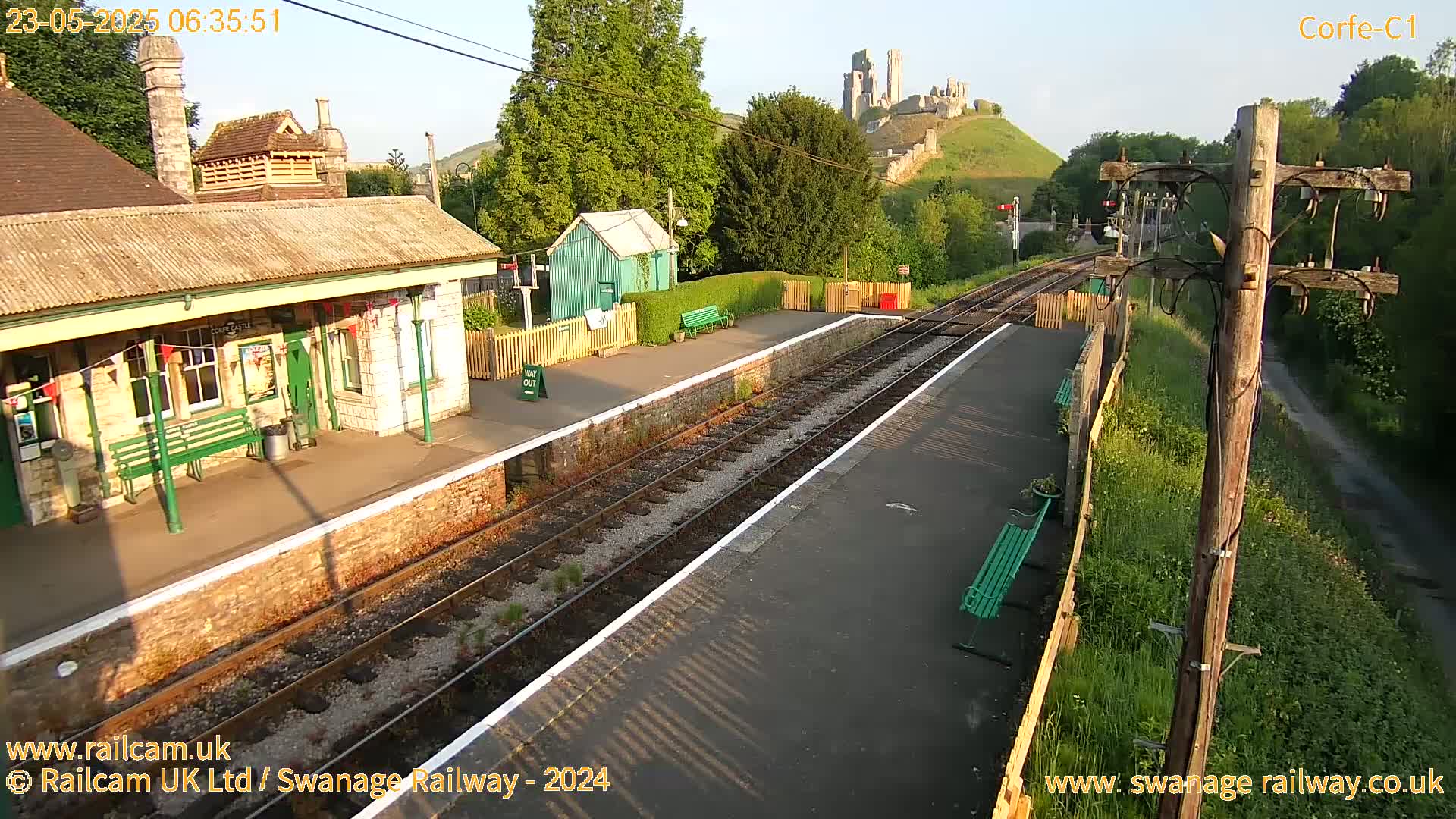 A train station platform with a stone building and a castle ruin visible on a hill in the background under a partly sunny sky.