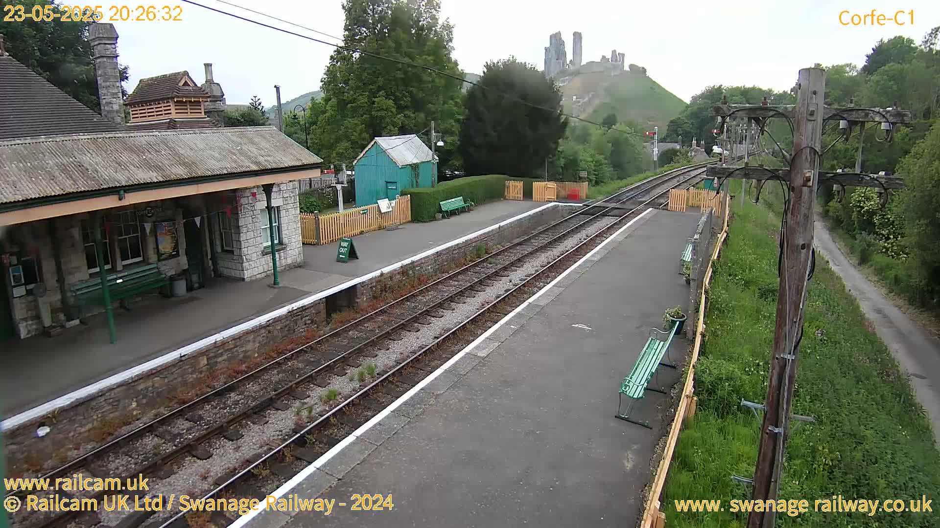 A train station platform with a stone building and benches, overlooking a hill with a castle ruin under an overcast sky.