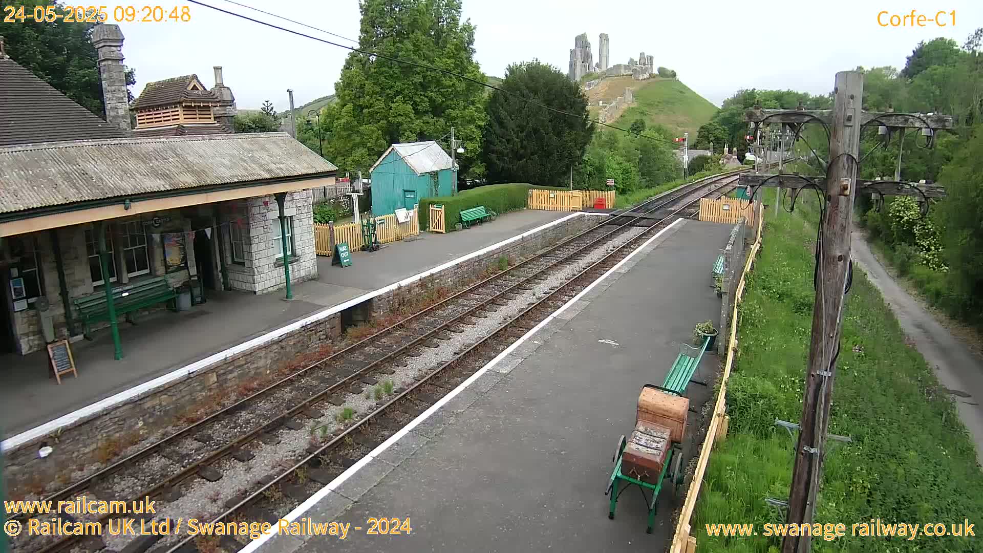 A train station platform with a stone building, a small green shed, and train tracks extends to a hilltop with a ruined castle in the background, under a partly cloudy sky.