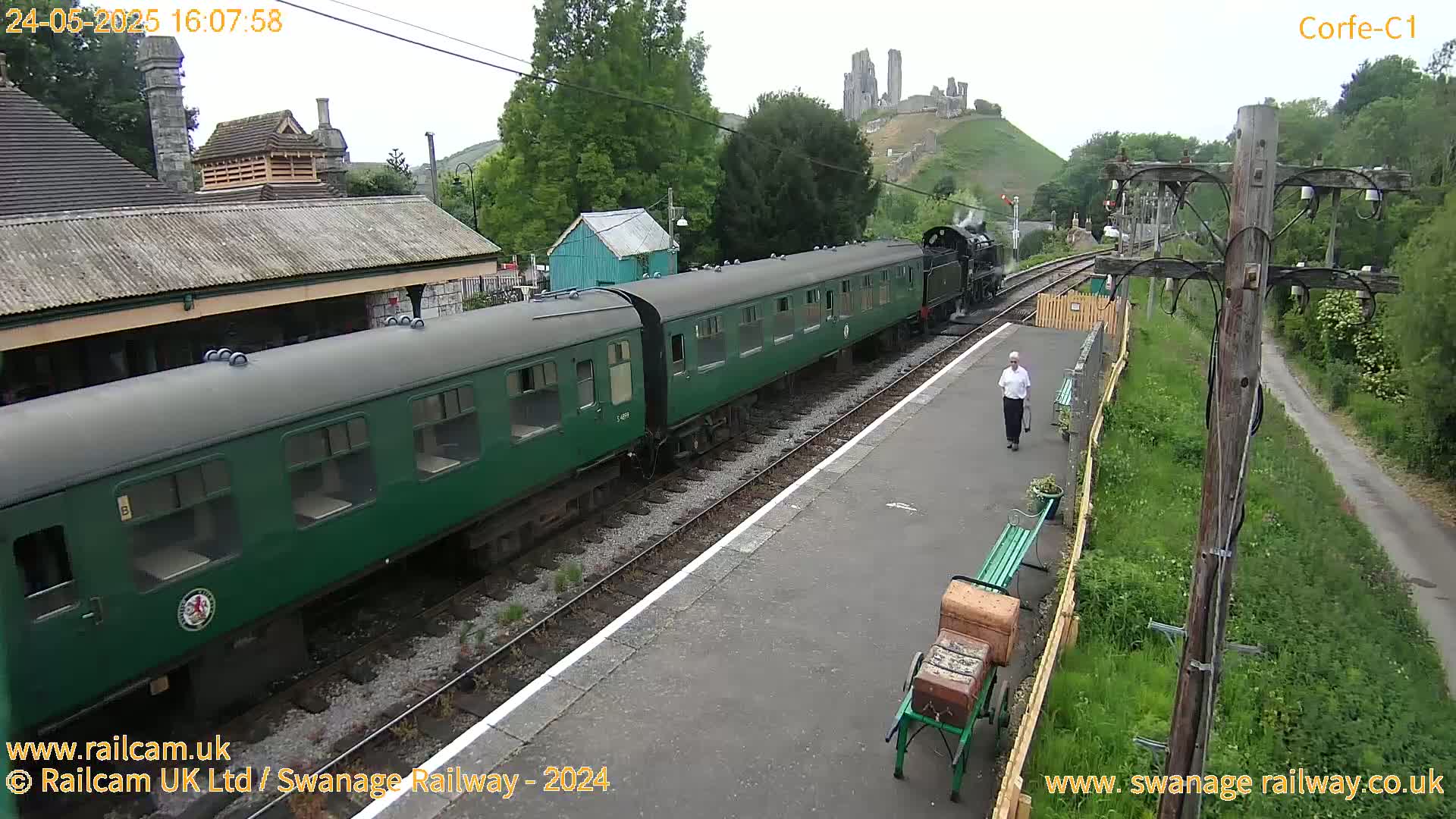 A green passenger train pulls into a station on an overcast day, with a person walking along the platform and a ruined castle visible in the distance.
