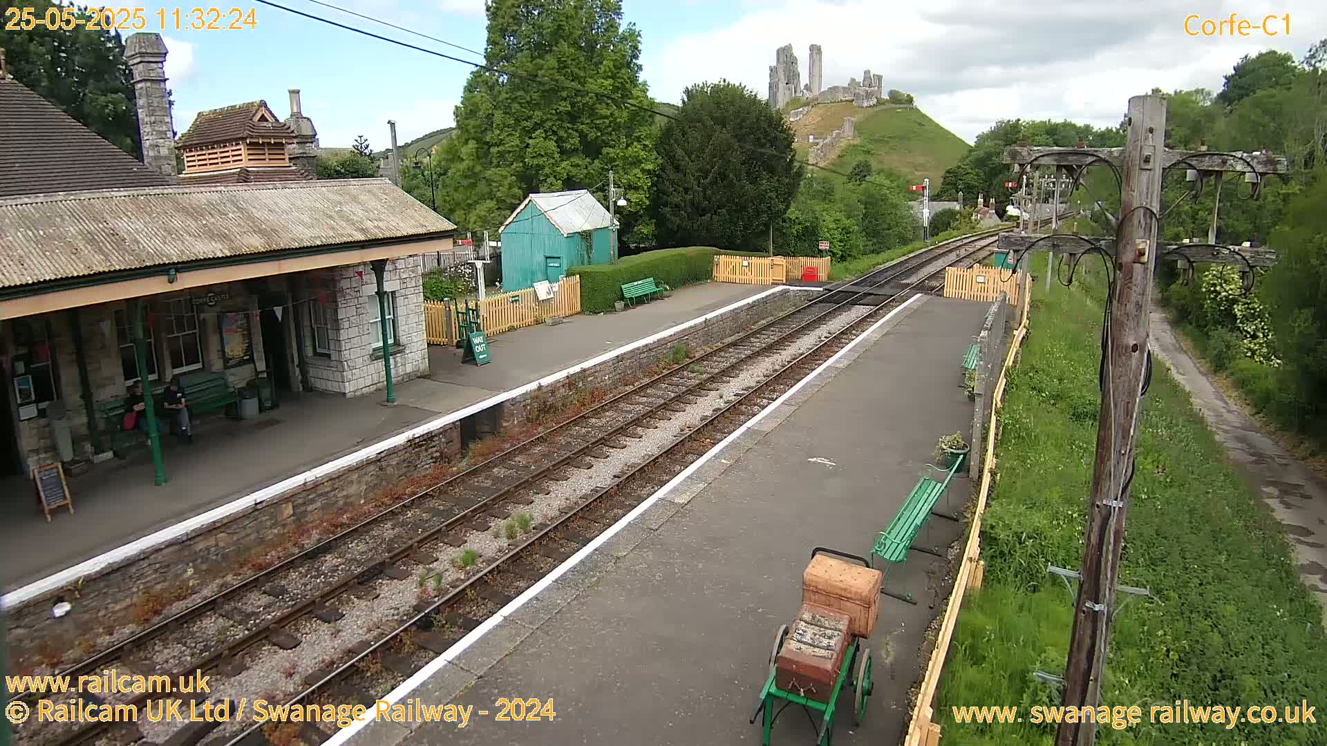 A train station with a stone building, a green shed, and train tracks, situated on a partly cloudy day with a hill and castle ruins visible in the background.