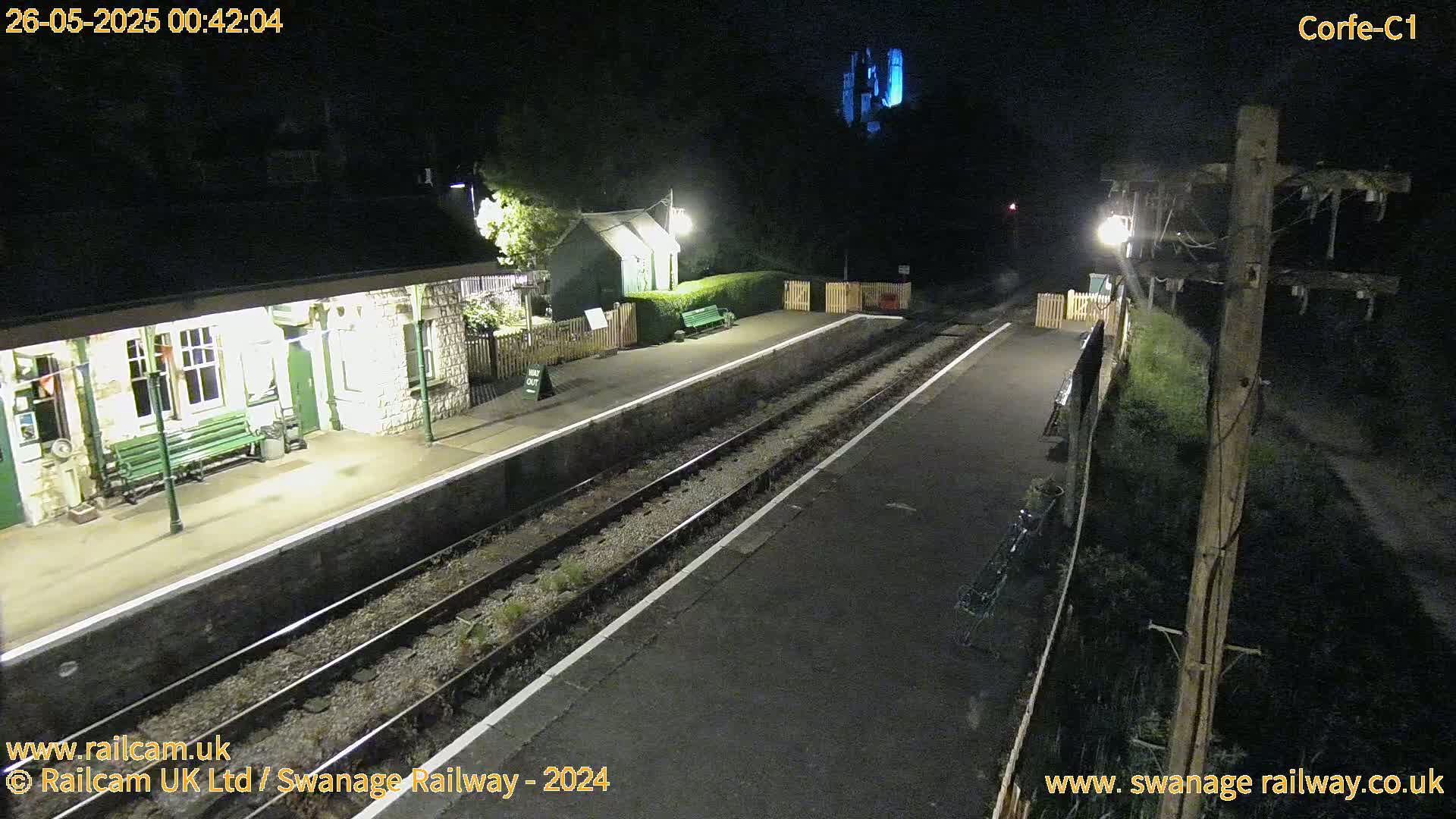 A nighttime view of a deserted train station platform with two sets of train tracks, a small building, and a partially visible structure in the background under dark conditions.