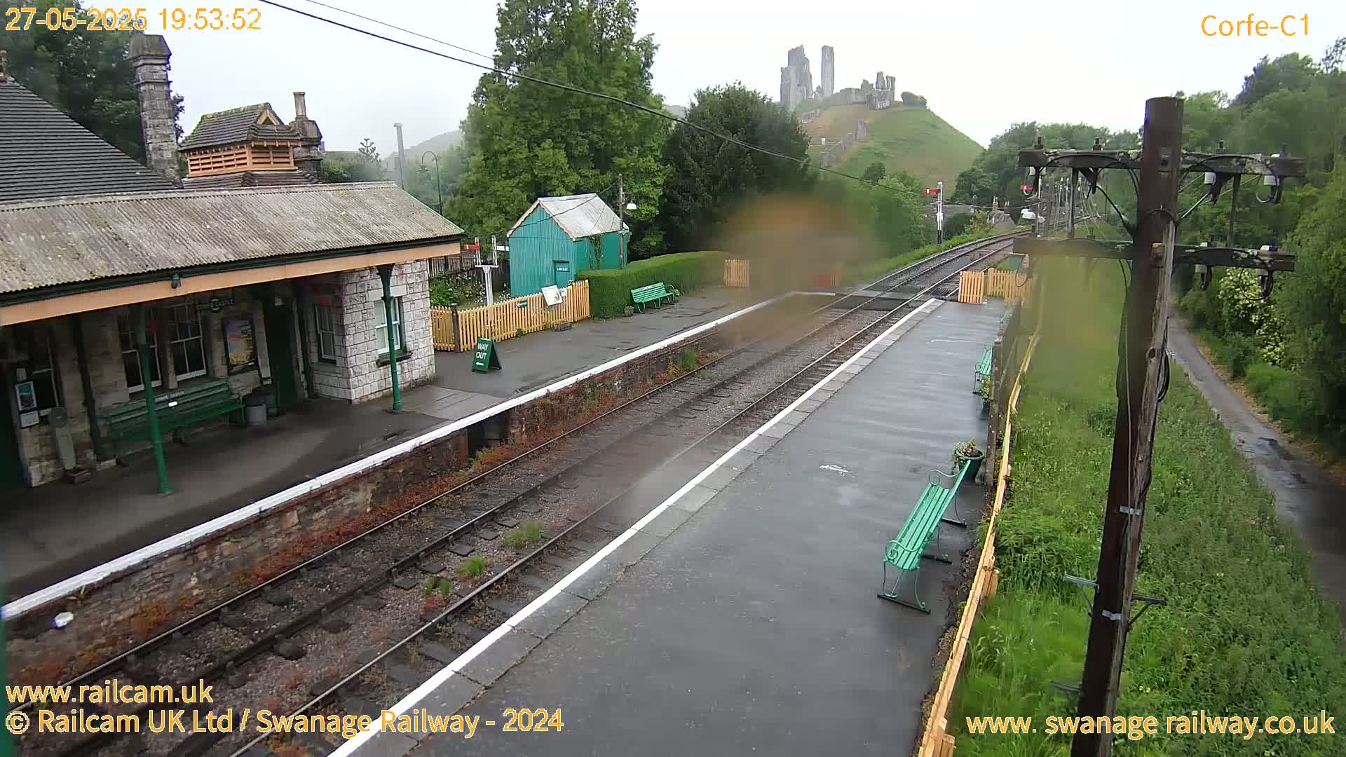 A train station with a stone building and benches, railway tracks, and a distant hilltop castle is seen on a rainy day.