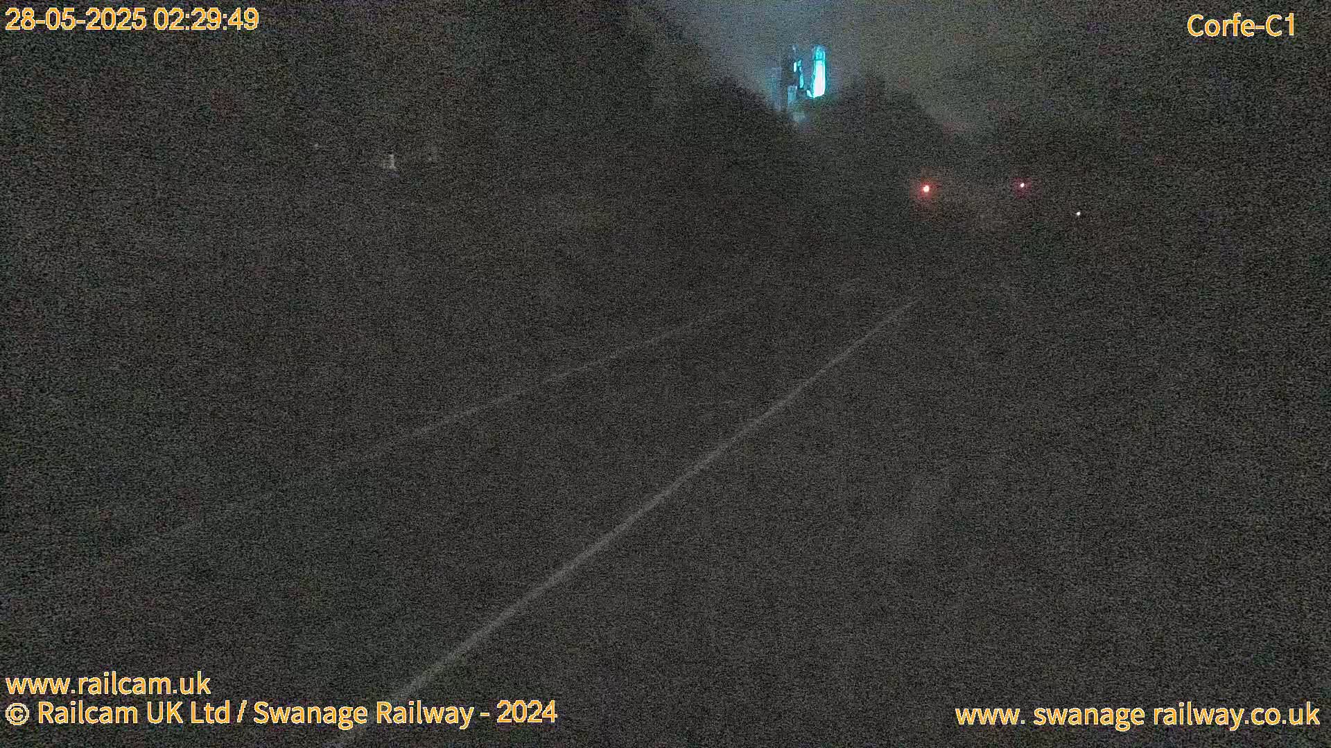 A dark, nighttime view of a railway track with faint white lines, and a few distant lights visible in the hazy darkness.