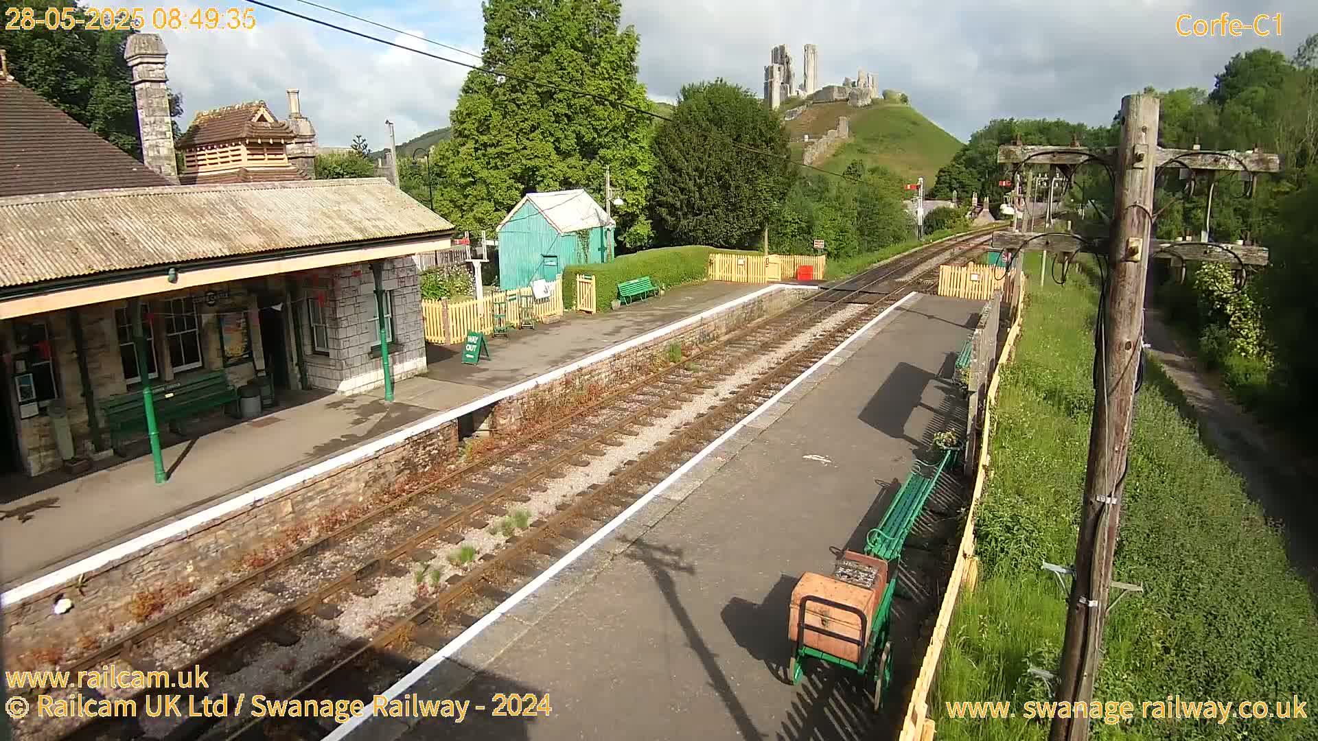 A railway station platform with a stone building and a small green shed, overlooking a hill with a ruined castle in the background, under a partly cloudy sky.