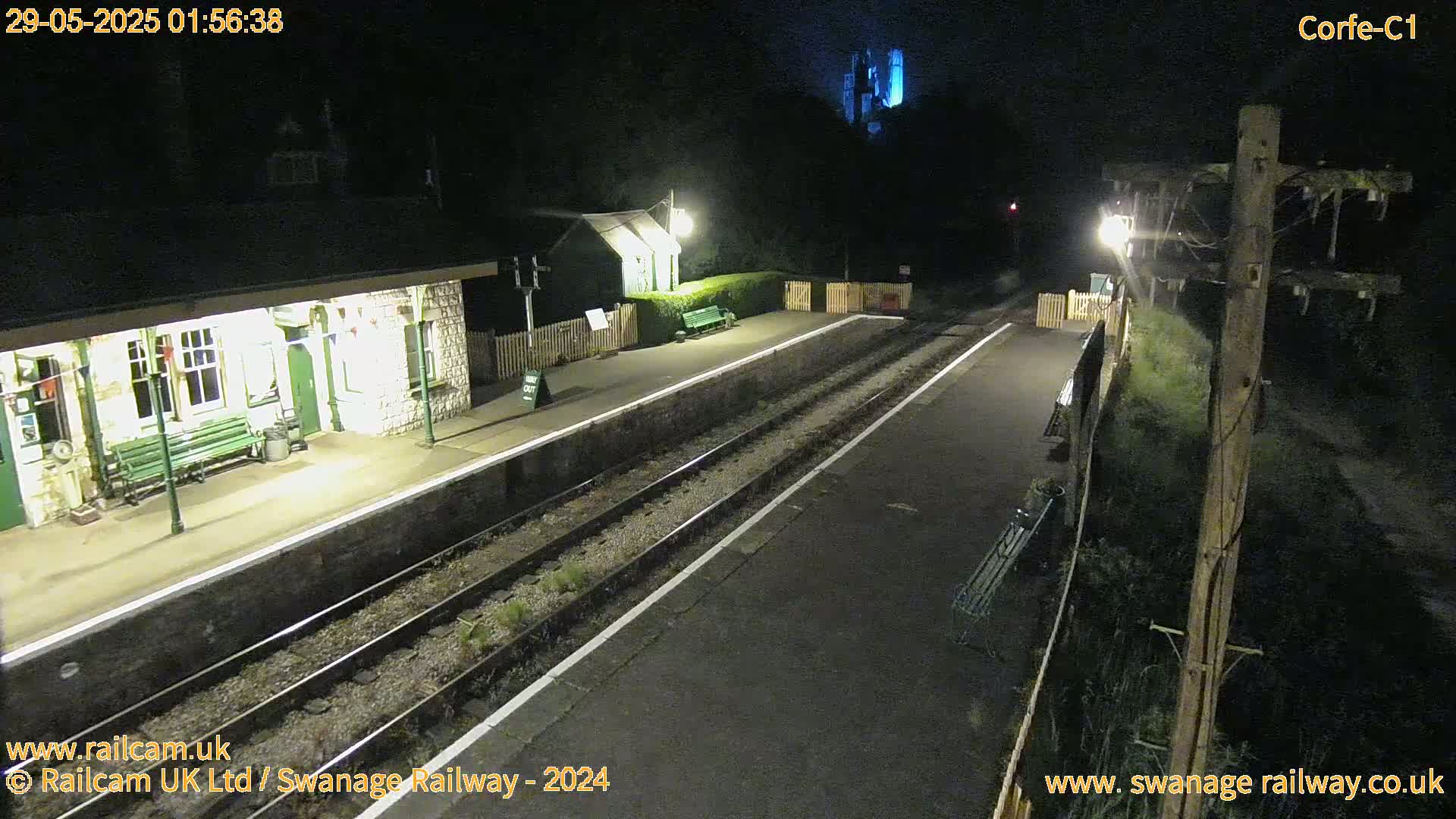 An empty train station platform at night is illuminated by lights, with train tracks extending into the darkness beyond.