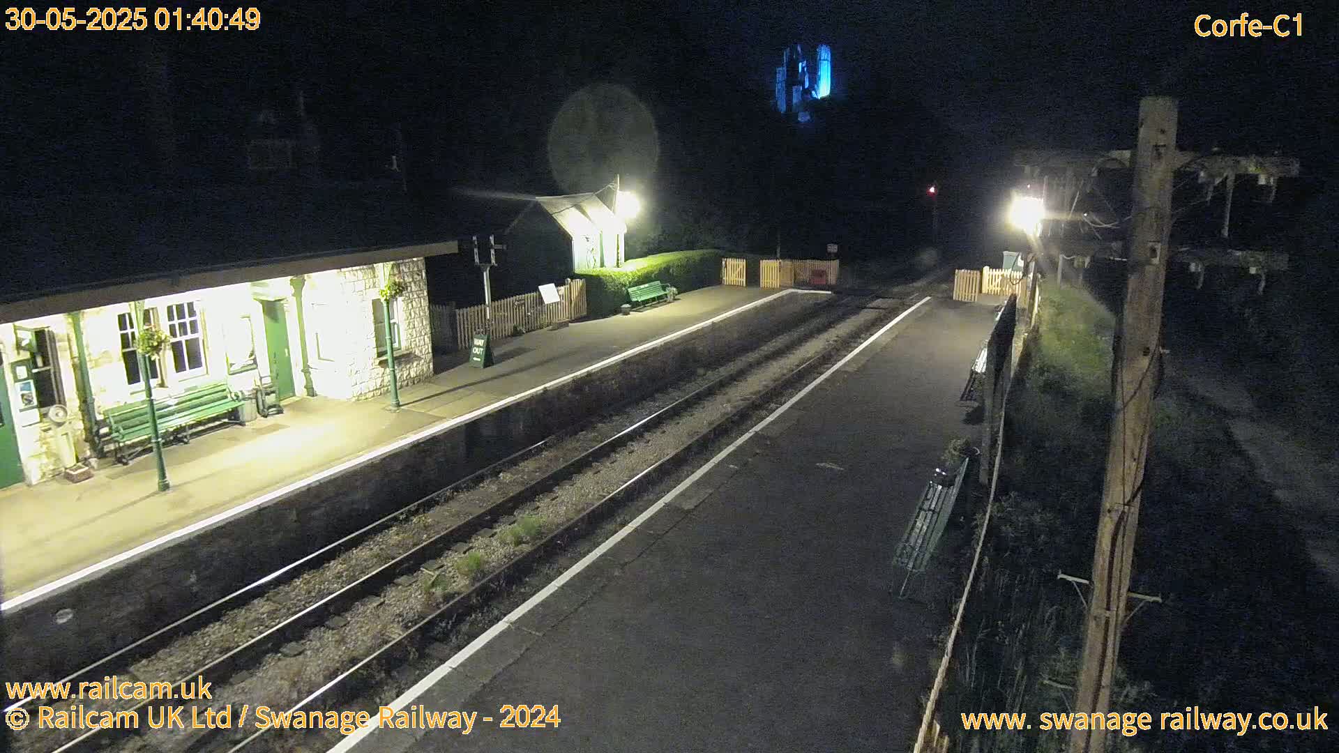 A nighttime view of a deserted train station platform with two sets of tracks, illuminated by artificial lights.