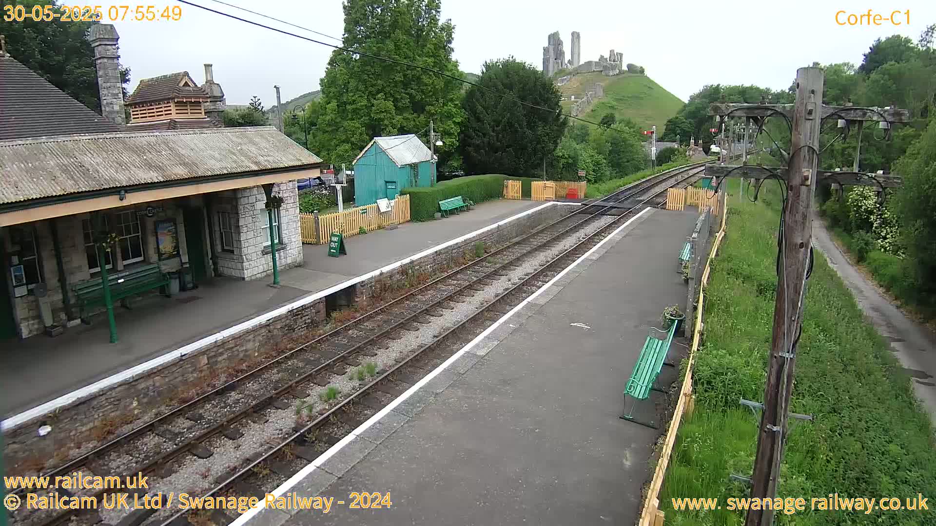 A train station platform with a stone building, a small green shed, and a distant ruined castle on a hill under an overcast sky.