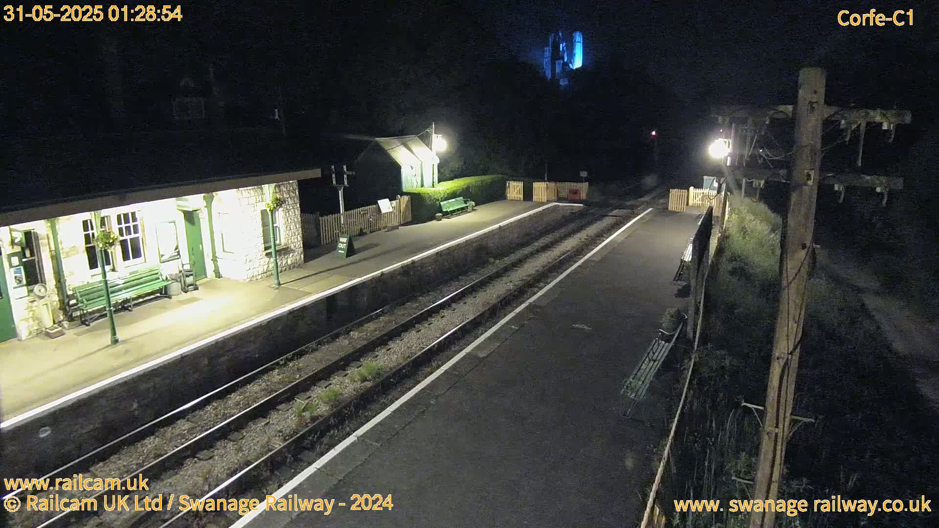 A nighttime view of a deserted train station platform with two parallel tracks, a small building, and some vegetation, illuminated by artificial light.