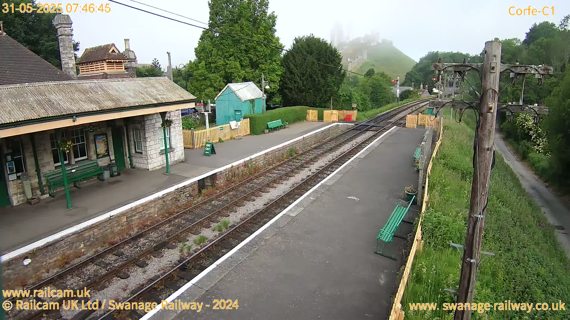 A train station with a stone building, a small green shed, and several benches sits on a railway line with a hill shrouded in fog in the background, under an overcast sky.