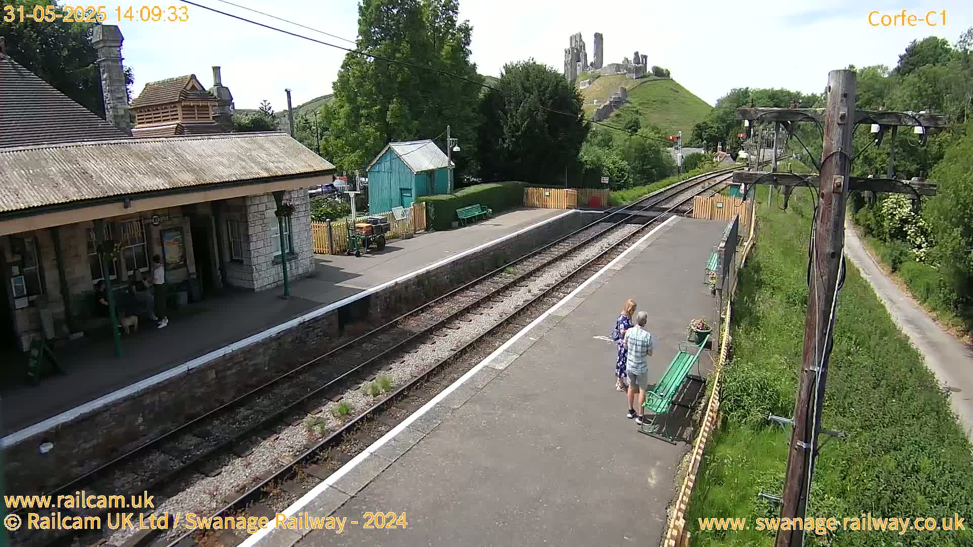 A train station platform on a sunny day, with a stone building, a small green shed, and a distant ruined castle visible on a hill.