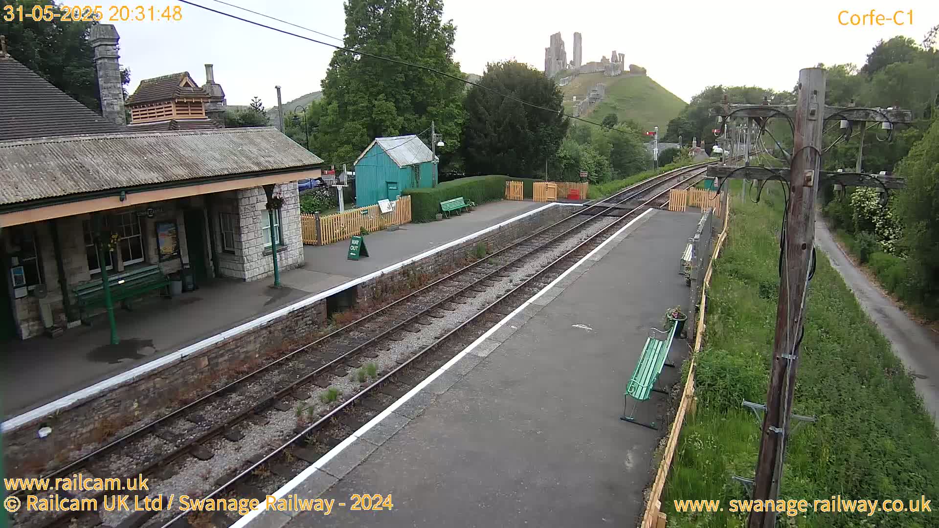 A train station platform with a stone building and benches, overlooking a hill with a castle ruin under a partly cloudy sky.