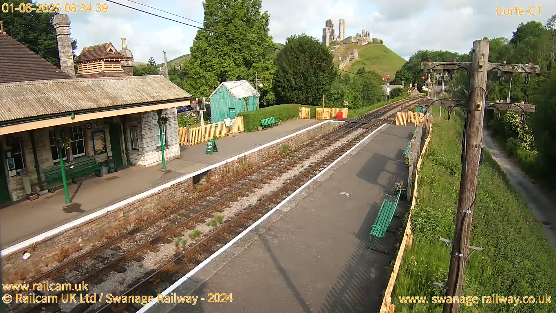 A train station platform with a stone building and a partially ruined castle visible in the distance on a partly sunny day.