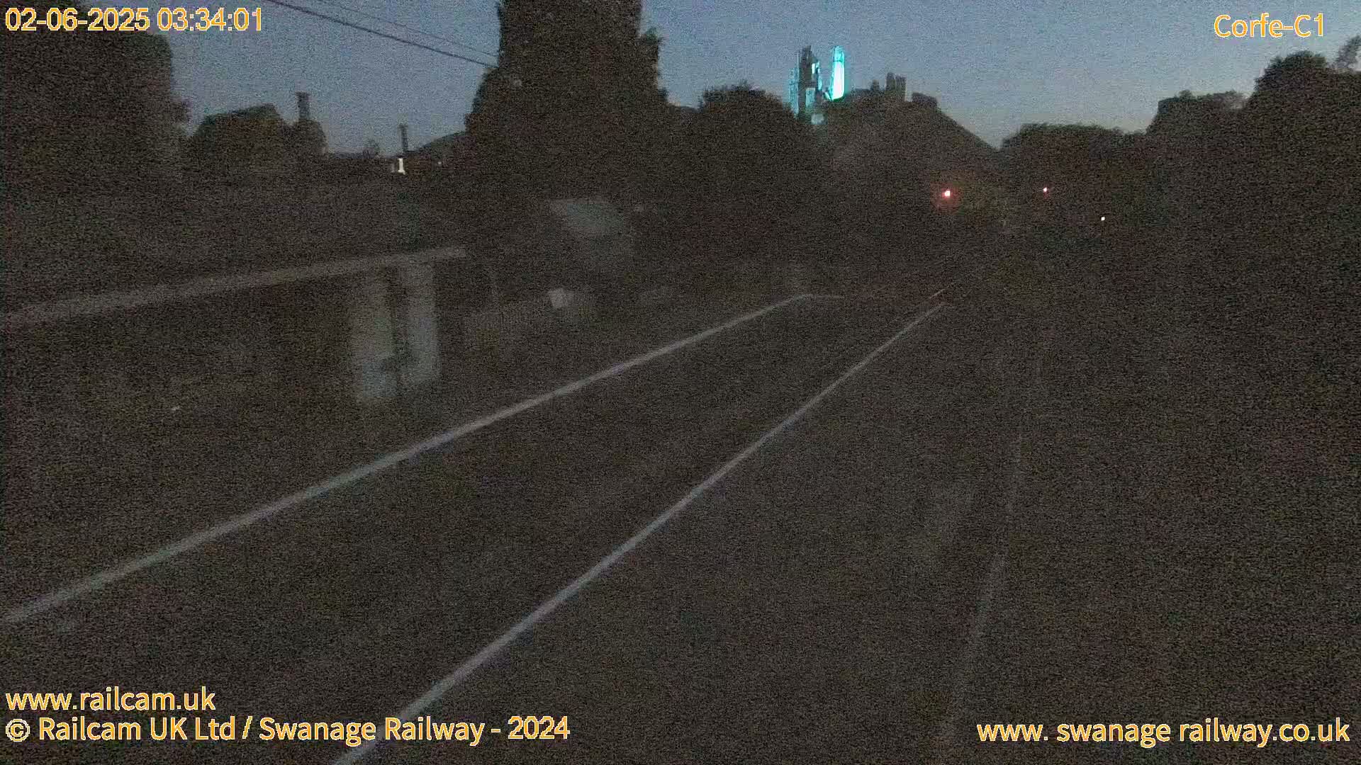 A dark, nighttime view of a railway track leading towards a dimly lit building and trees under a twilight sky.