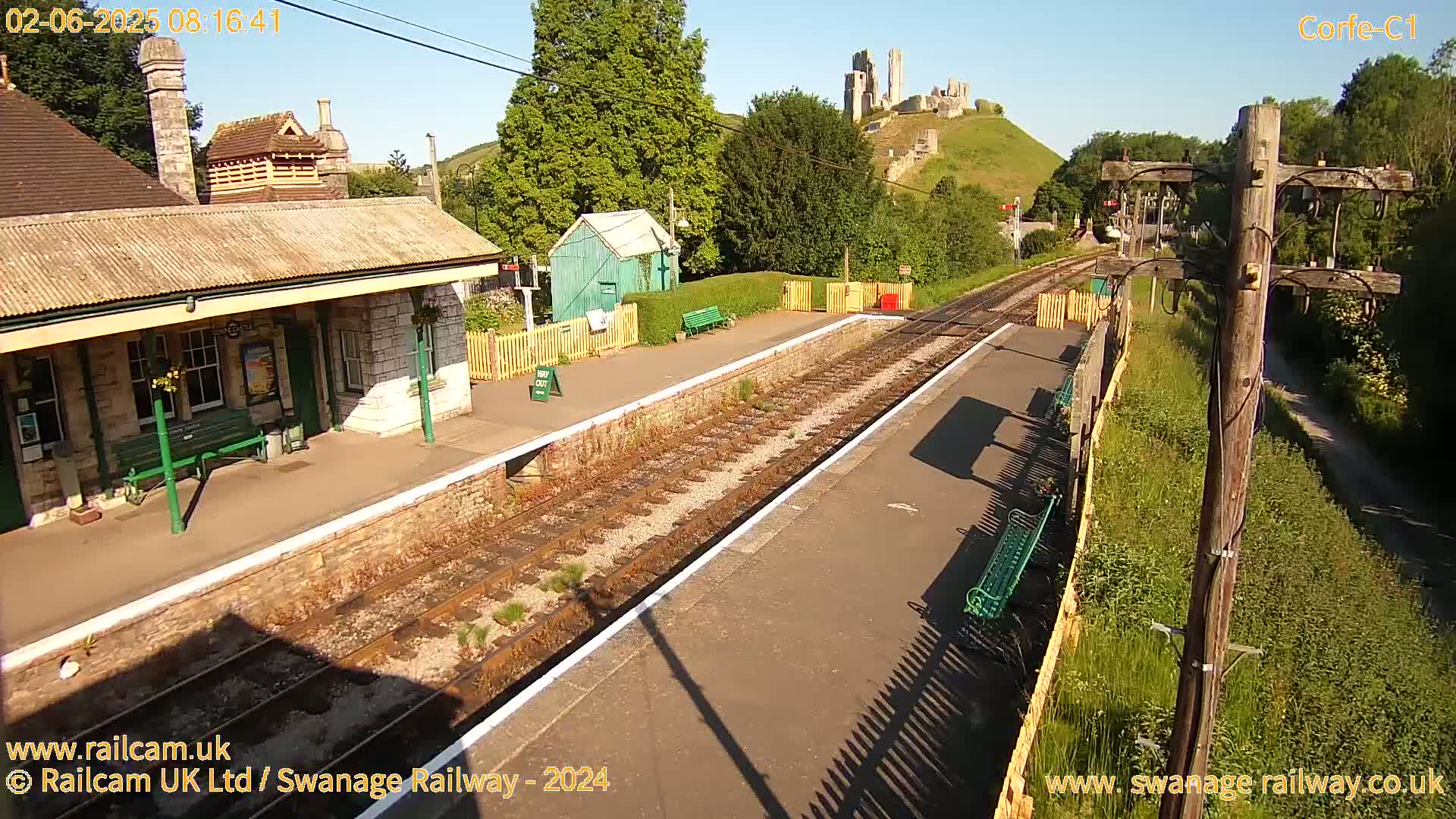 A train station platform with a stone building, a small green shed, and a castle visible in the distance on a sunny day.