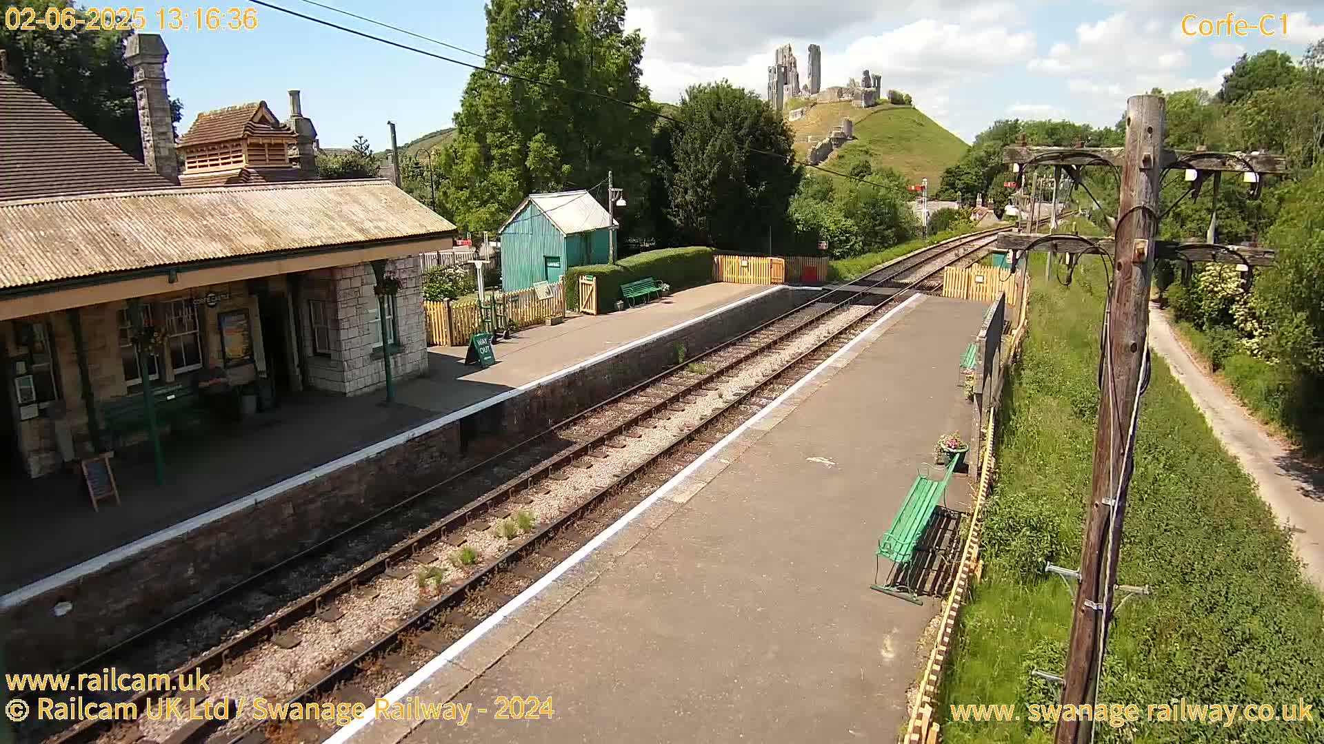 A train station platform with two tracks, a stone station building, and a small green shed, overlooks a hill with a partially ruined castle under a sunny sky.