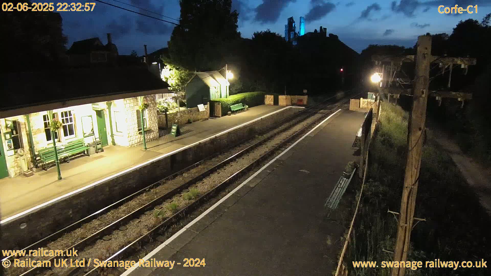 A deserted train station at night, illuminated by artificial lights, with a castle visible in the distance under a dark sky.