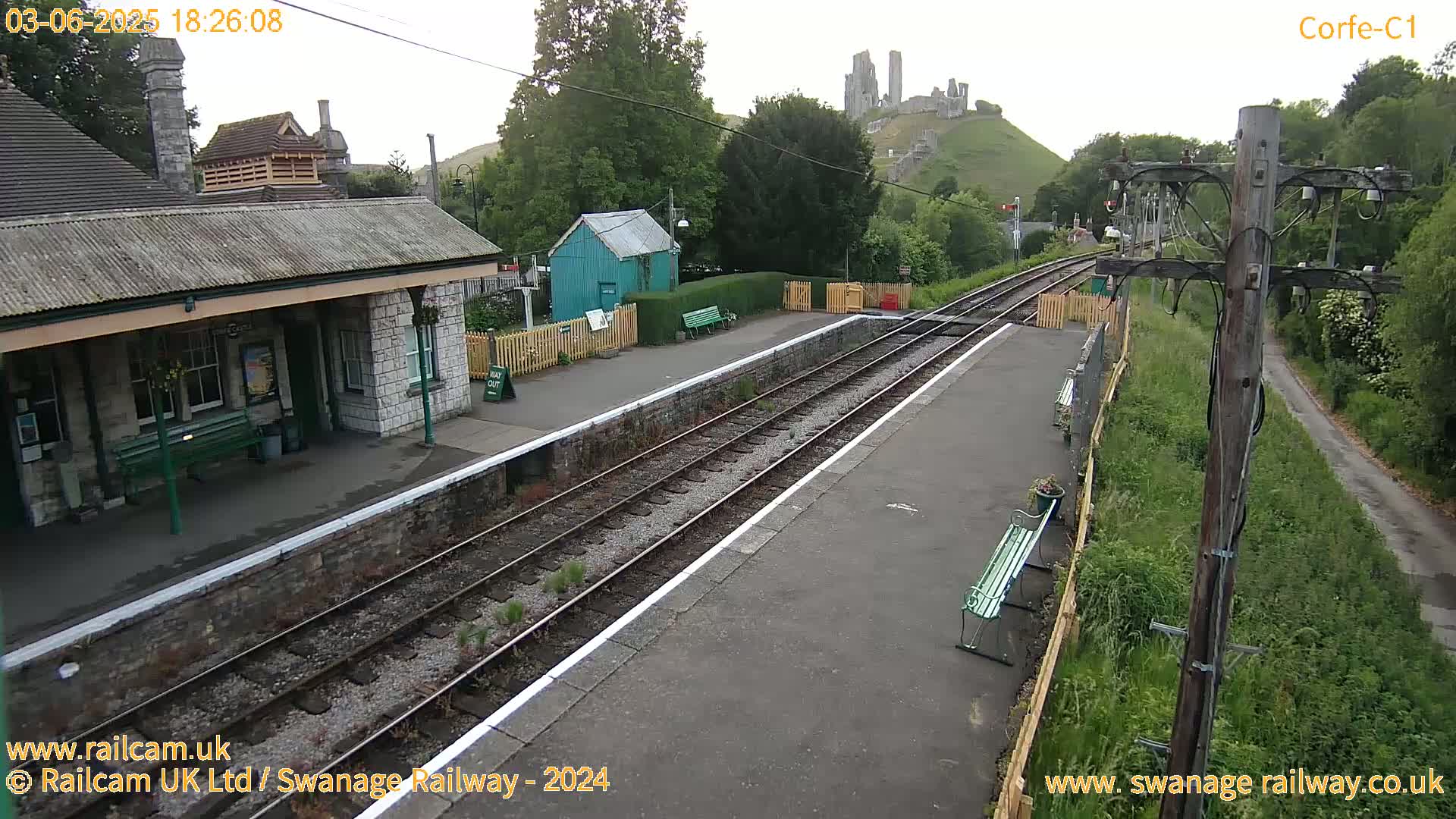 A train station platform with a stone building, a small green shed, and a distant hilltop castle is visible under an overcast sky.