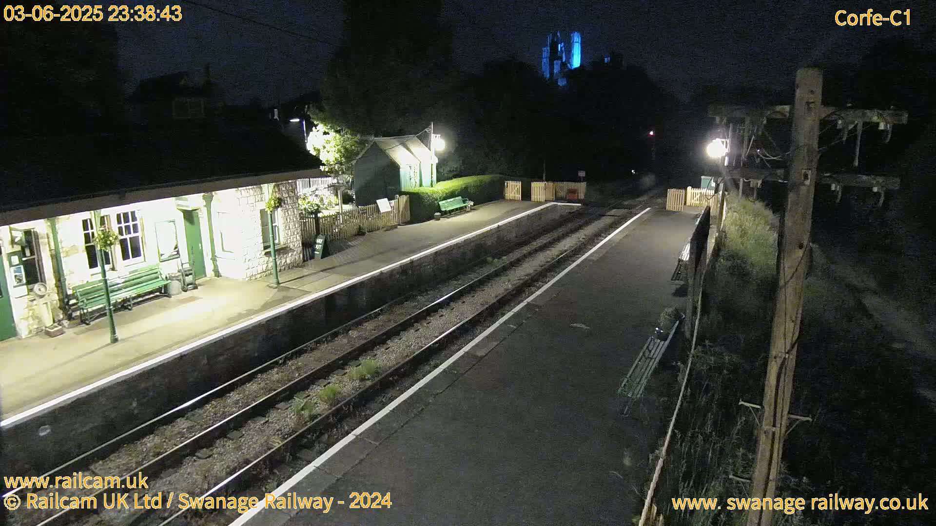 A nighttime view of a deserted train station platform with two sets of tracks, a stone building, and a dimly lit area beyond.