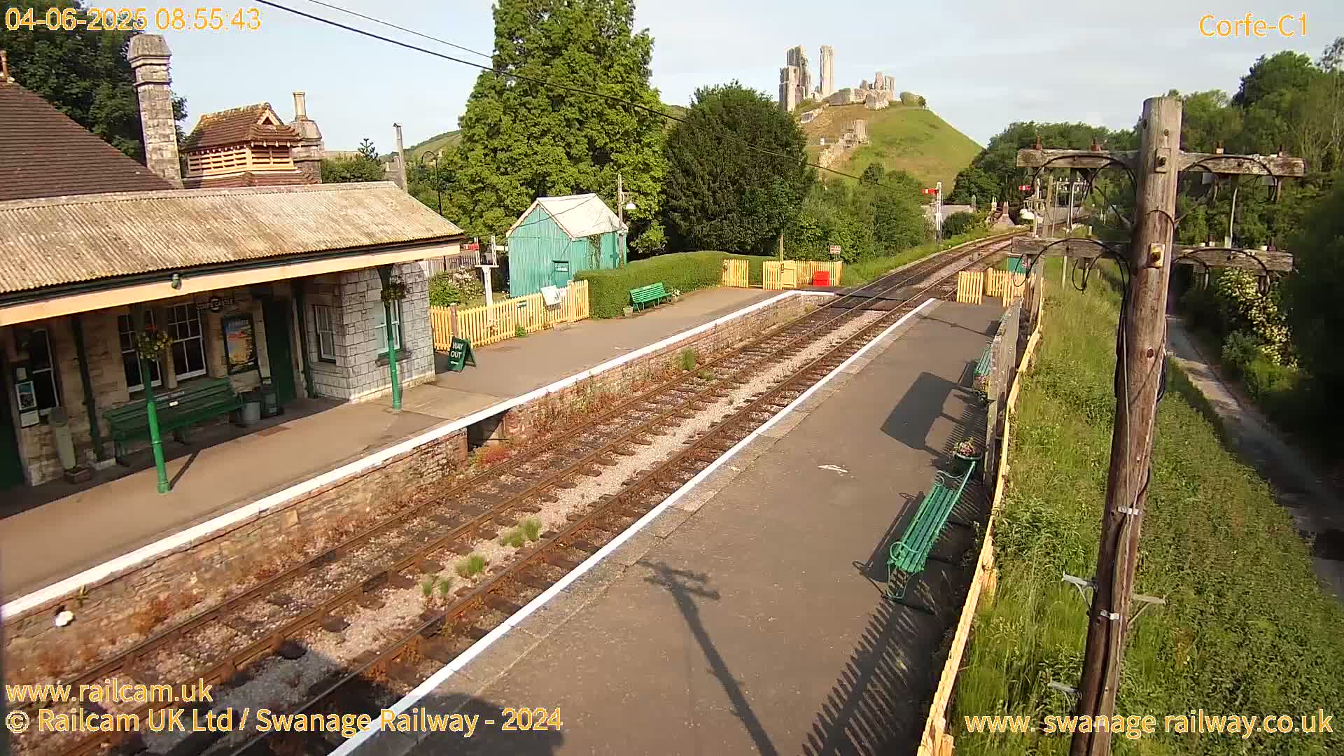 A train station platform with a stone building, a small green shed, and a hilltop castle in the background on a sunny day.