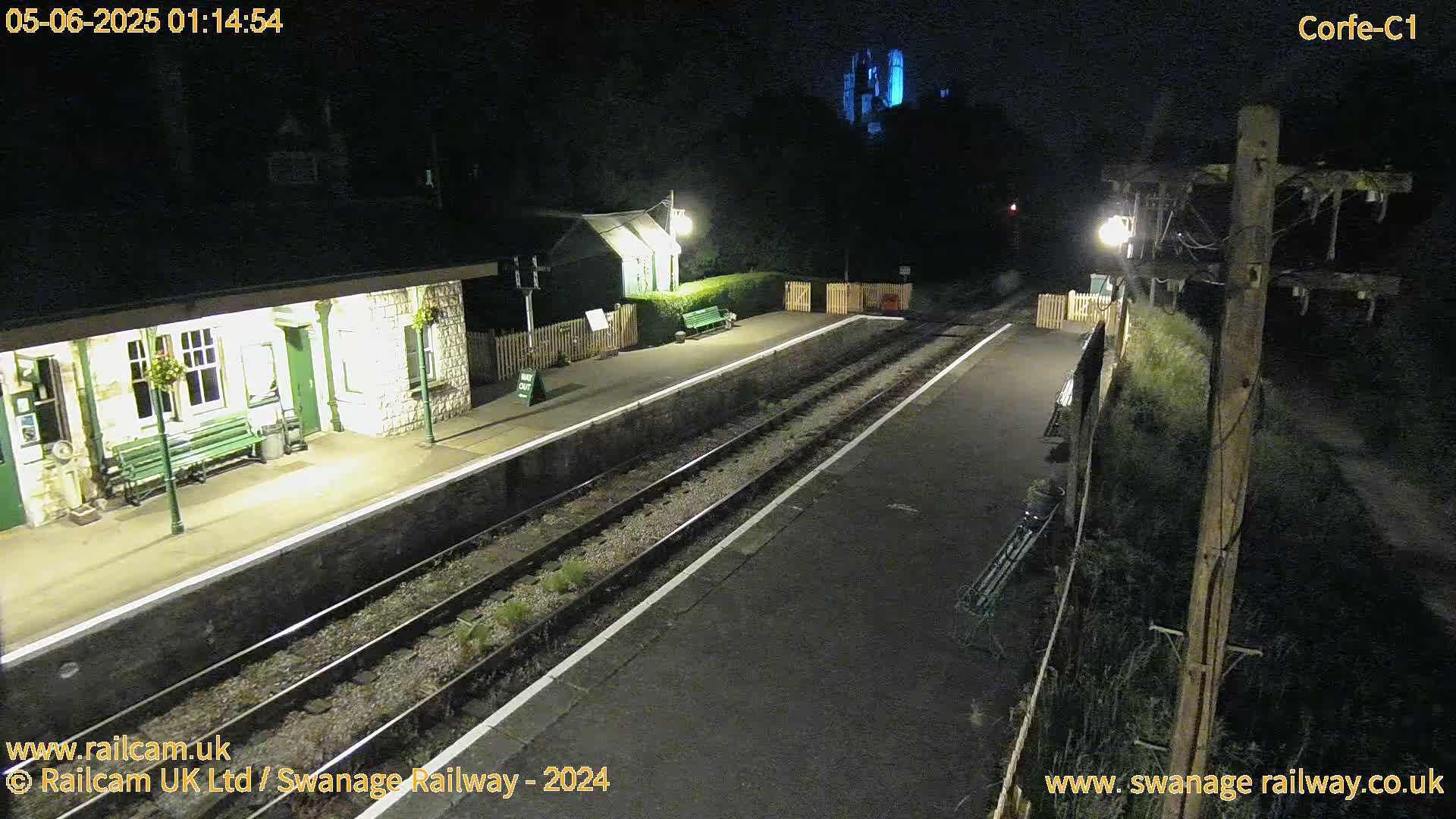 A nighttime view of a deserted train station platform with two sets of train tracks, a stone building, and a dimly lit area beyond.