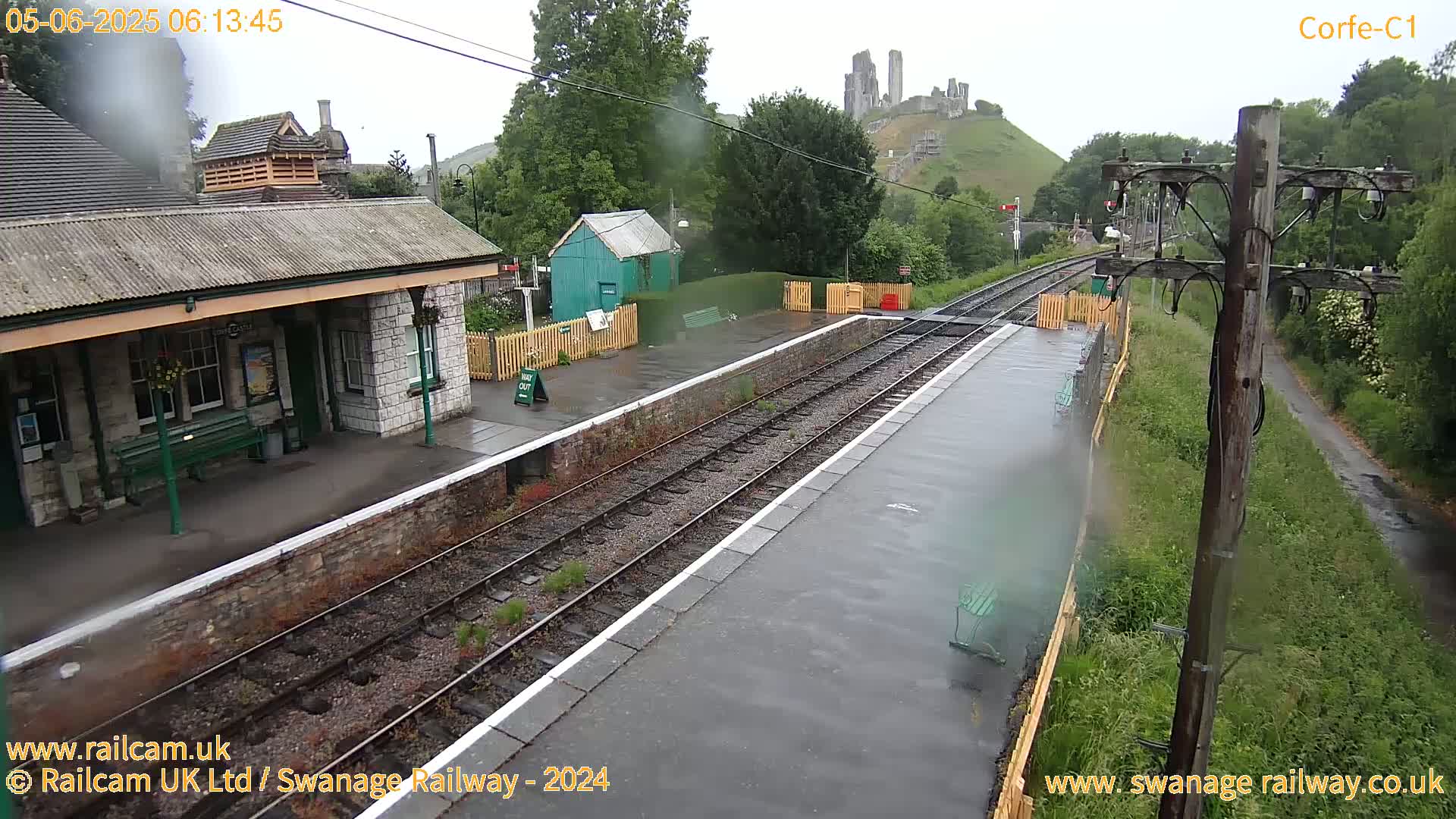 A wet train station platform with multiple train tracks, a stone station building, and a distant view of a hilltop castle under an overcast sky.