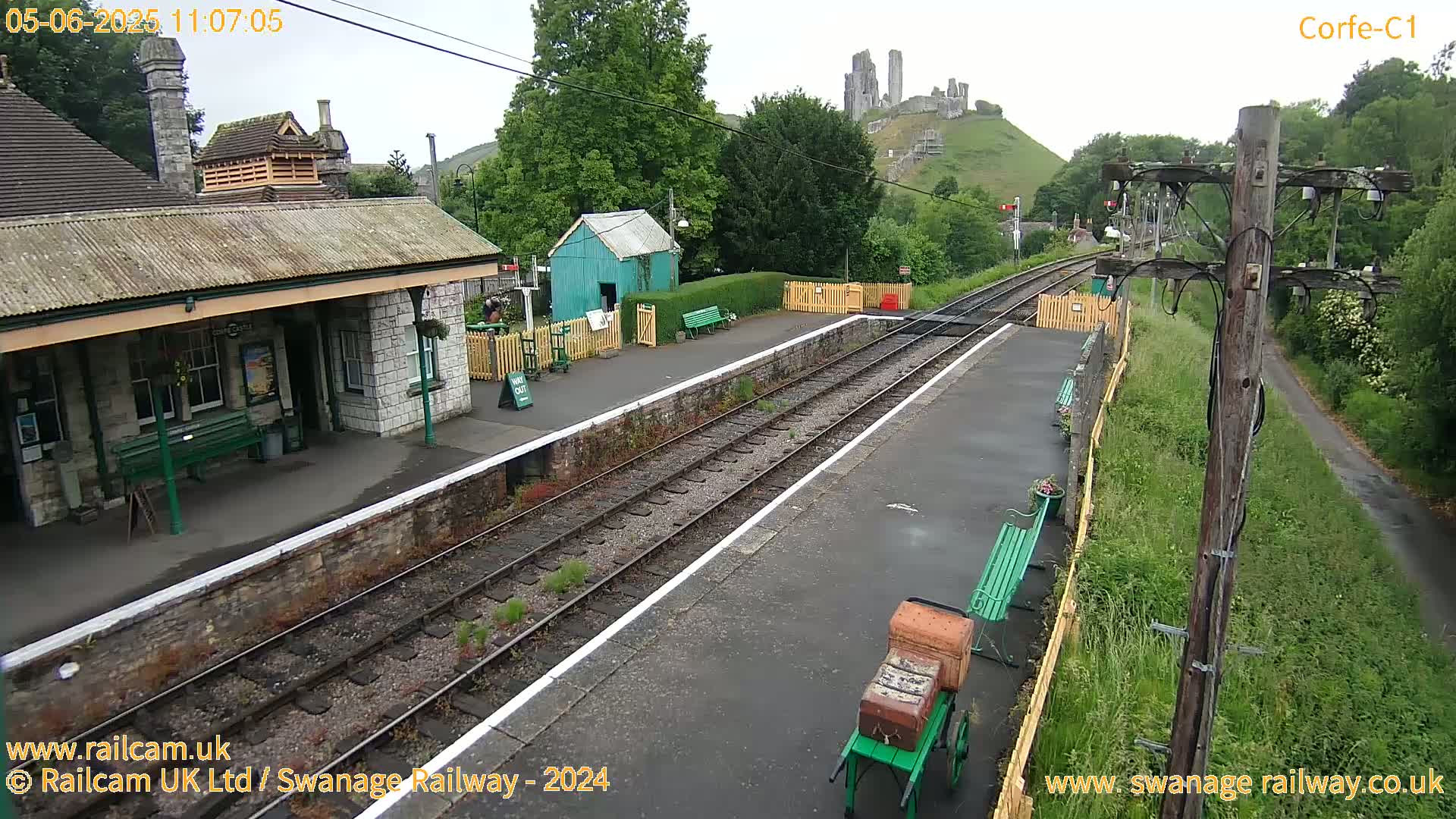 A train station platform with a stone building, a small green shed, and a distant castle is visible on a partly cloudy day.