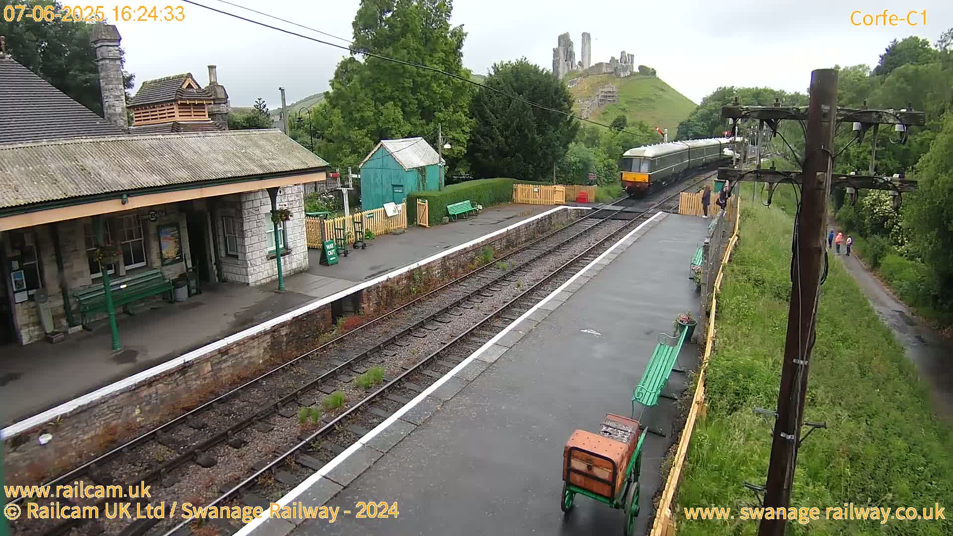 A train is at a railway station on an overcast day, with a stone ruin visible on a hill in the background.
