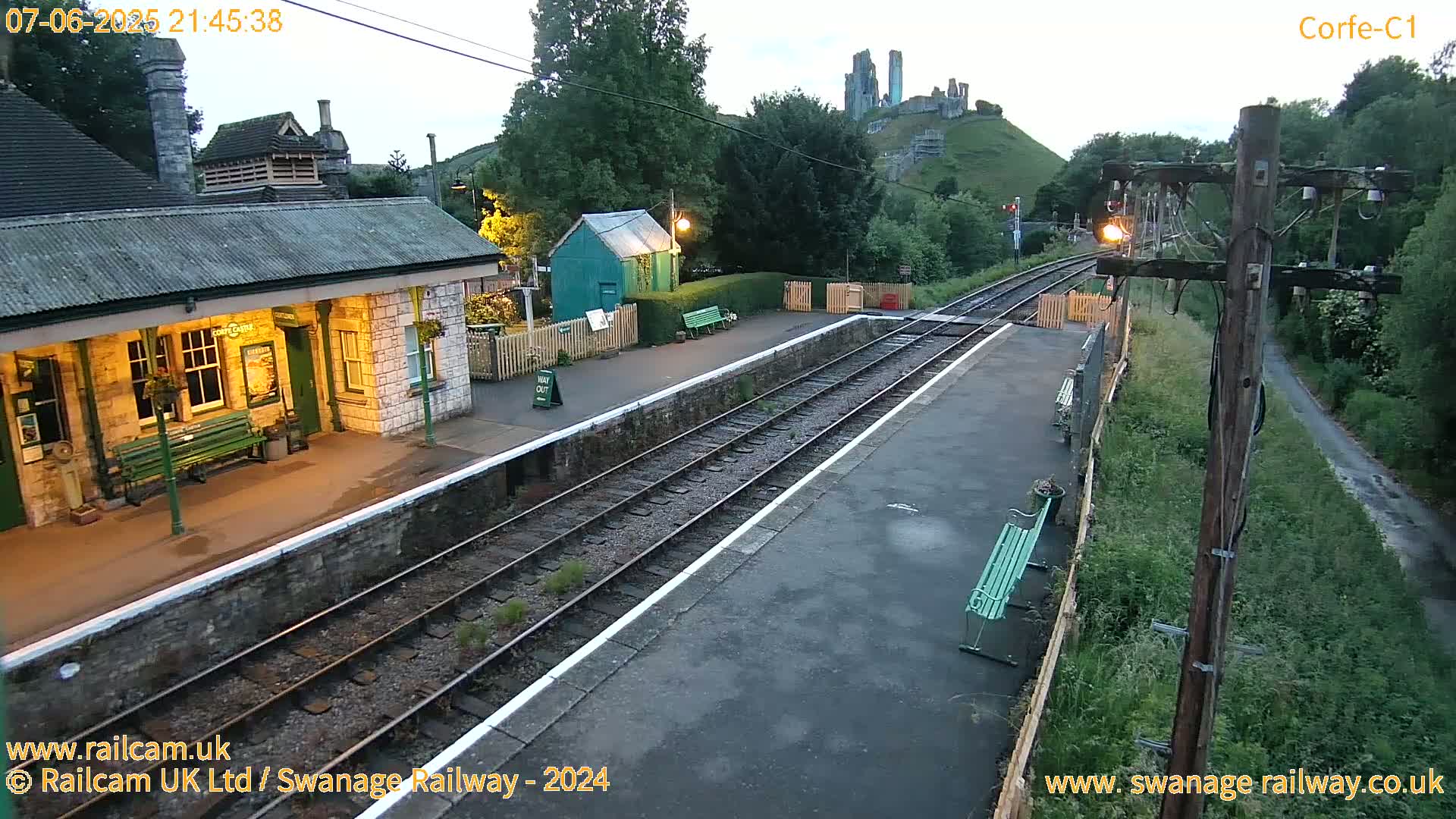 A train station at dusk, with a stone building and tracks, overlooks a hill with a partially ruined castle in the background.
