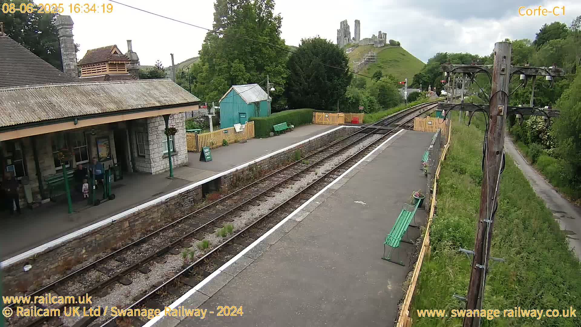 A train station platform with a stone building, a small green shed, and a hill with a castle ruin in the background under overcast skies.