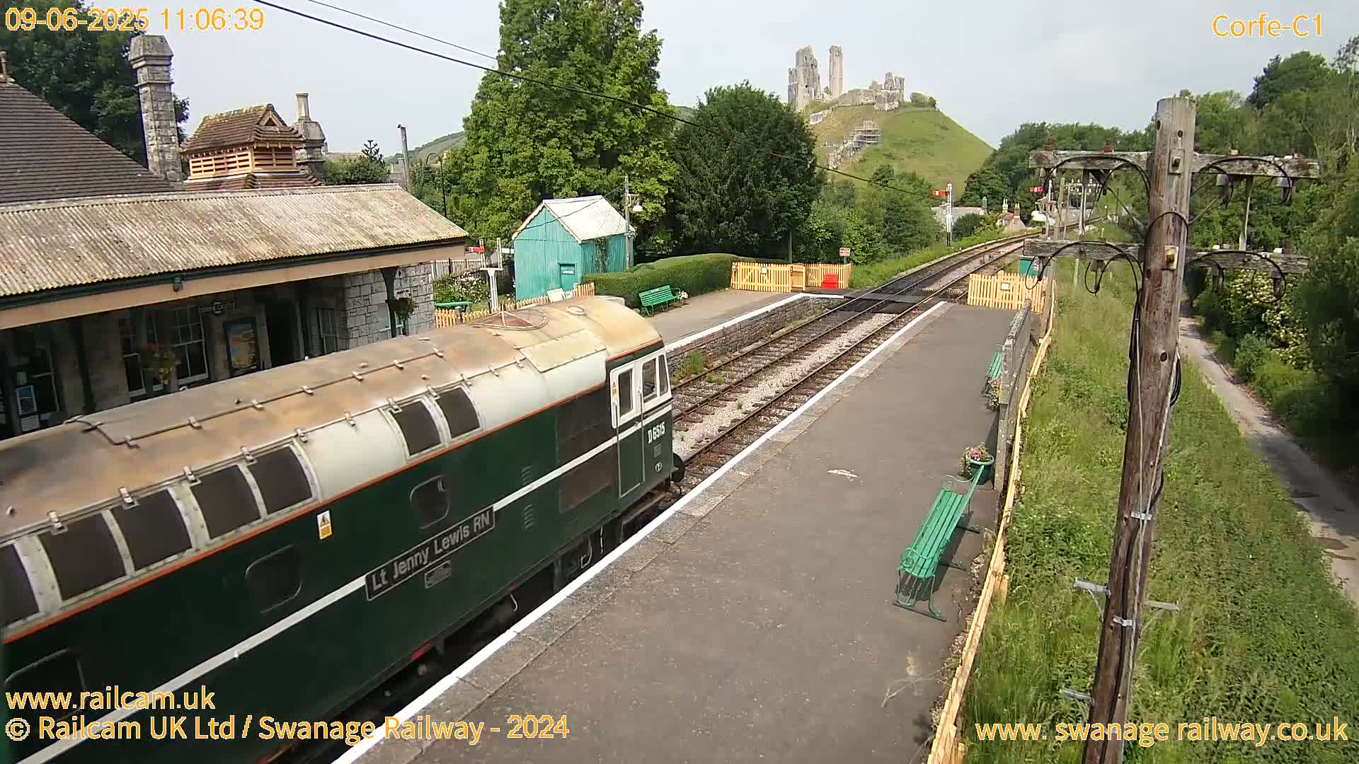 A green diesel train is arriving at a railway station on a partly sunny day, with a hilltop castle visible in the background.