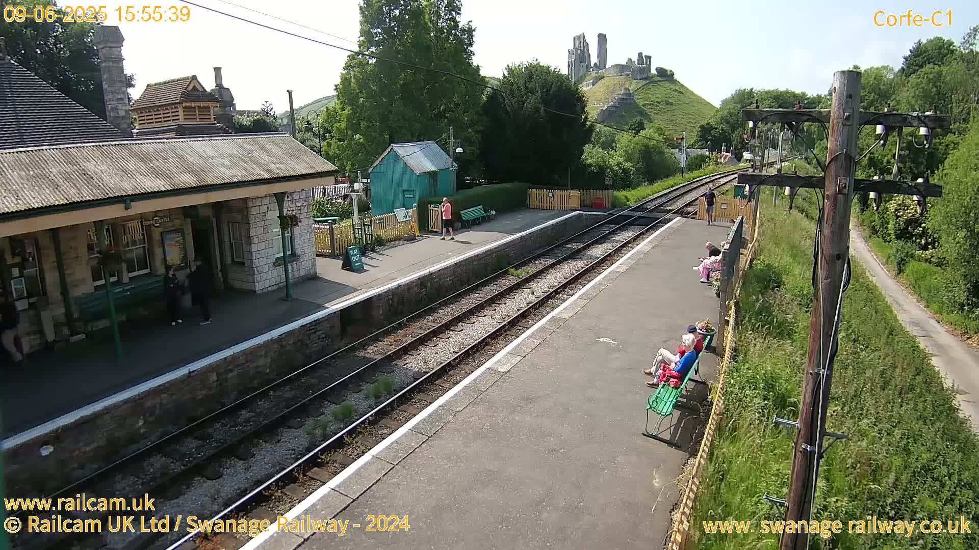 A train station platform with several people sitting on benches, a small building, and a castle ruin visible in the distance on a sunny day.