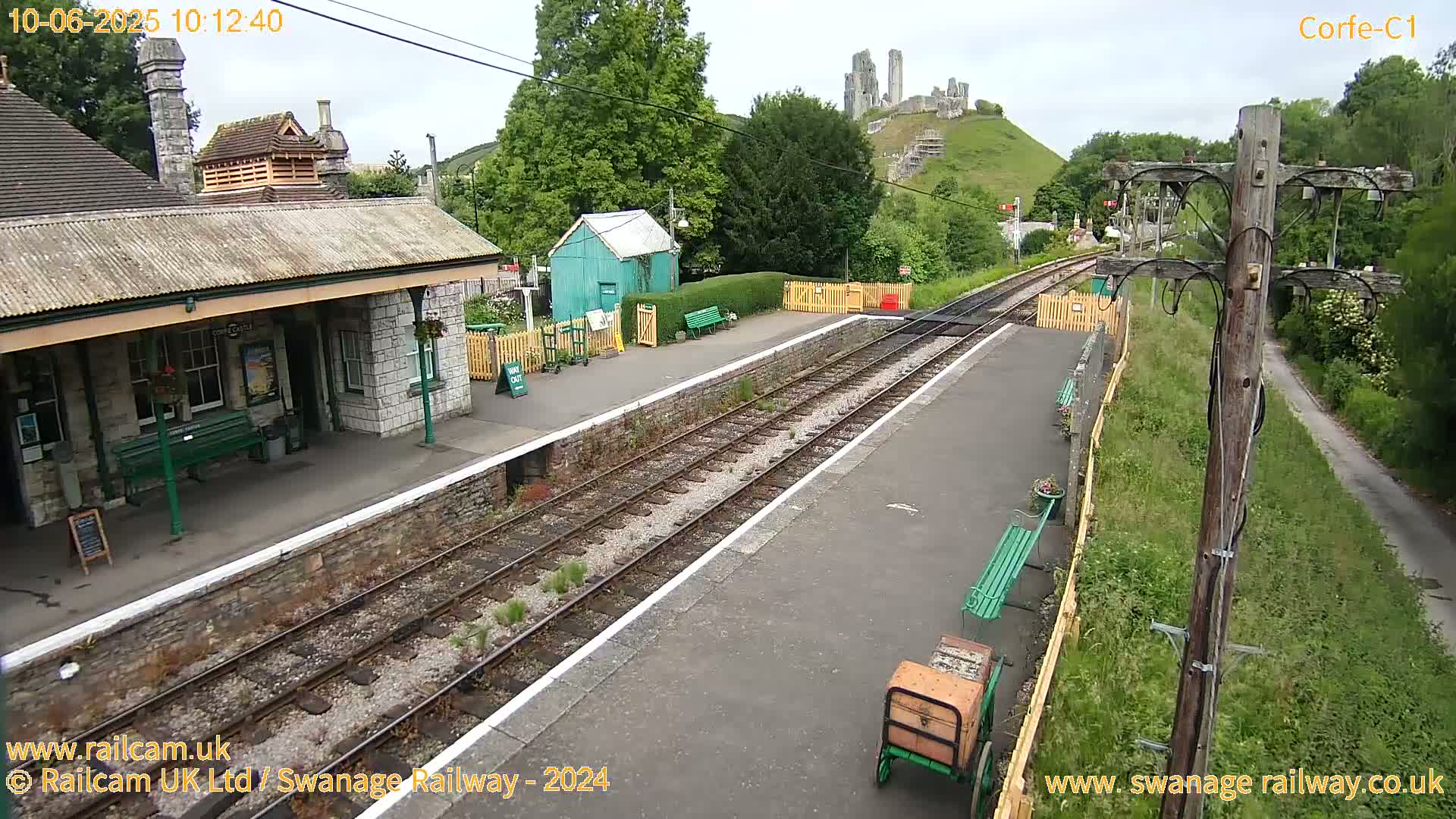 A train station platform with a stone building and a small green shed, situated under a partly cloudy sky, with a hill and ruins visible in the background.