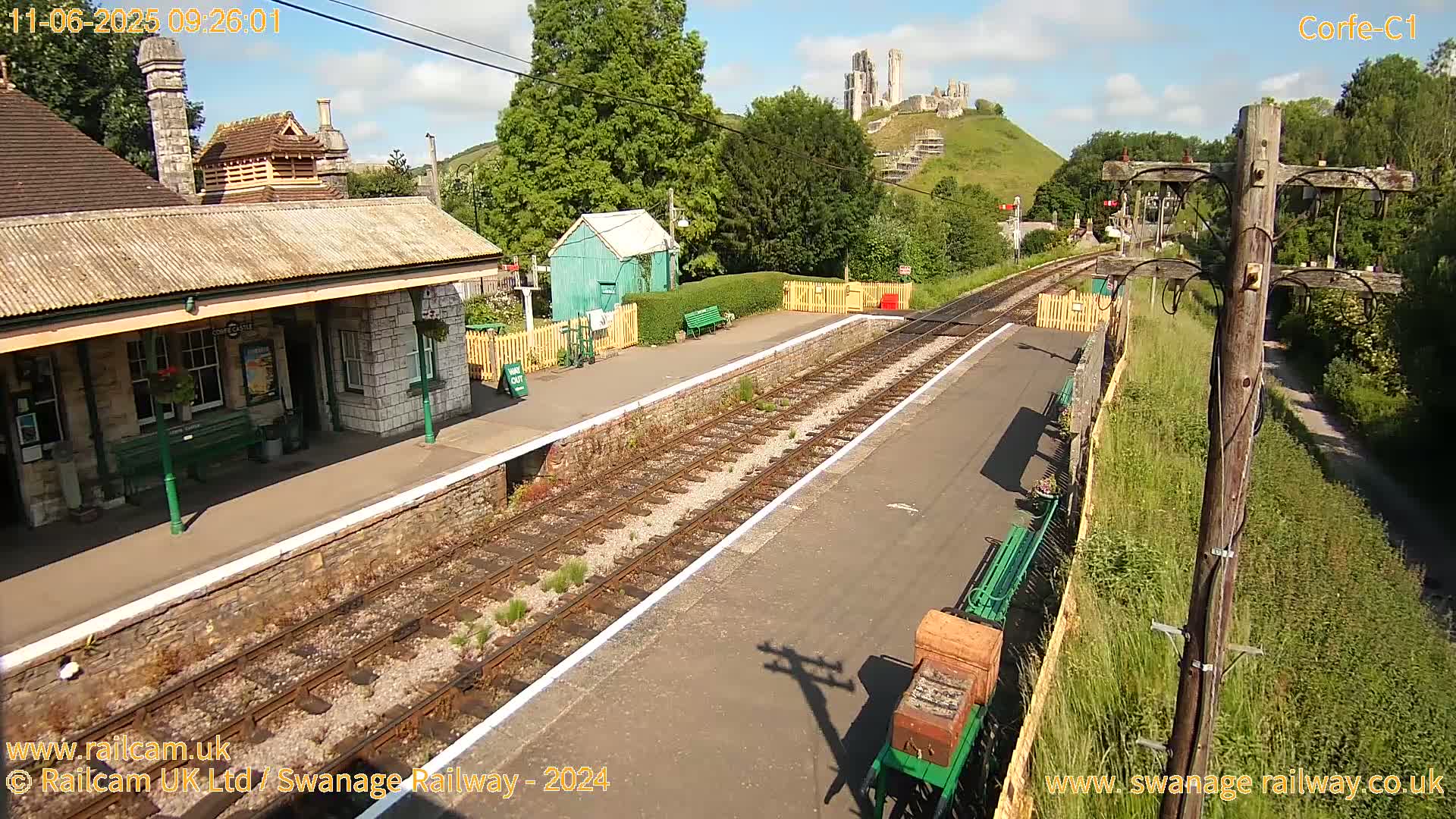 A sunny view of a train station platform with a stone building, a small green shed, and a hilltop castle ruin in the background.