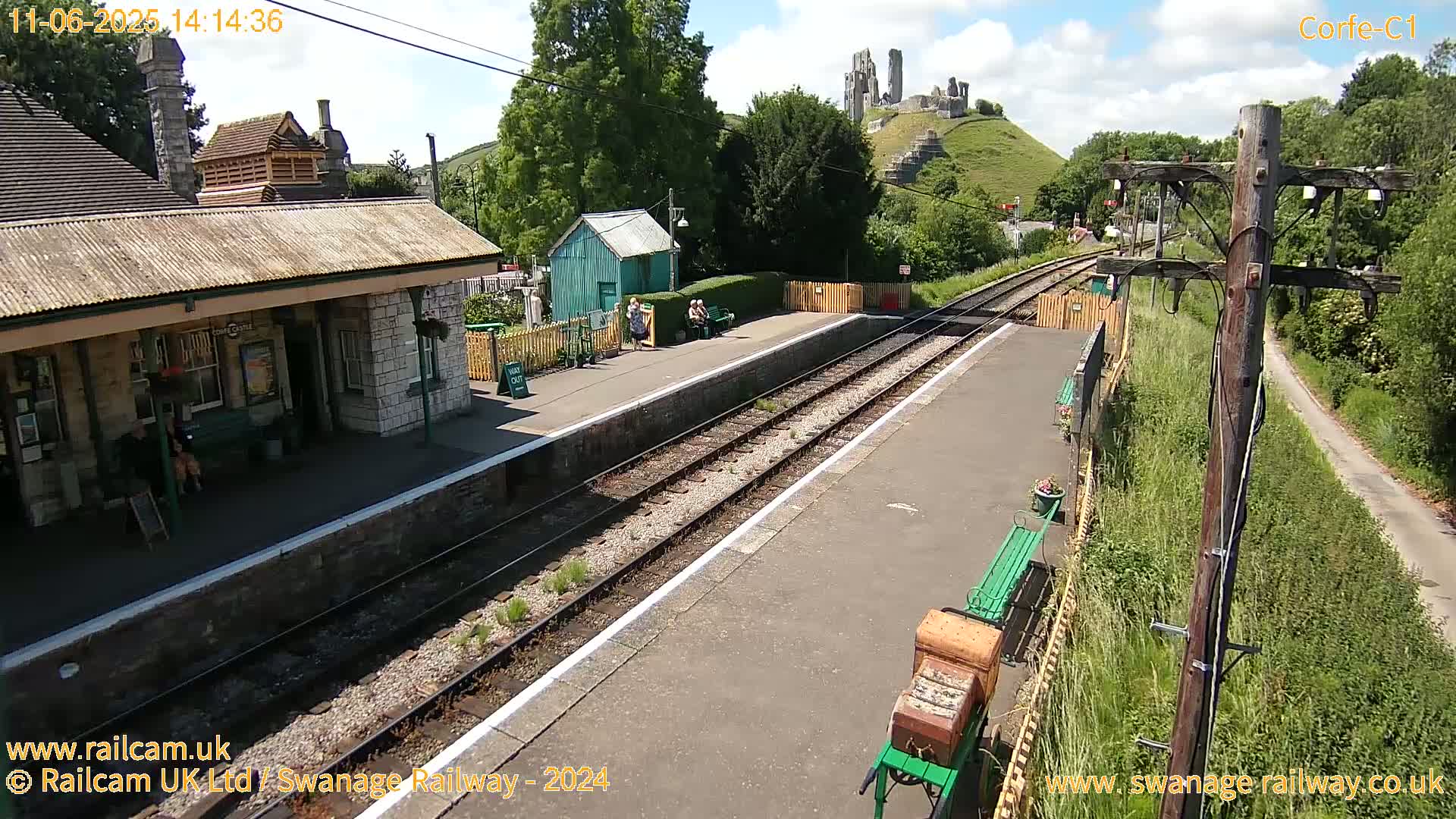 A train station platform with a stone building, a green shed, and a few people, under a sunny sky, with a hilltop ruin visible in the distance.