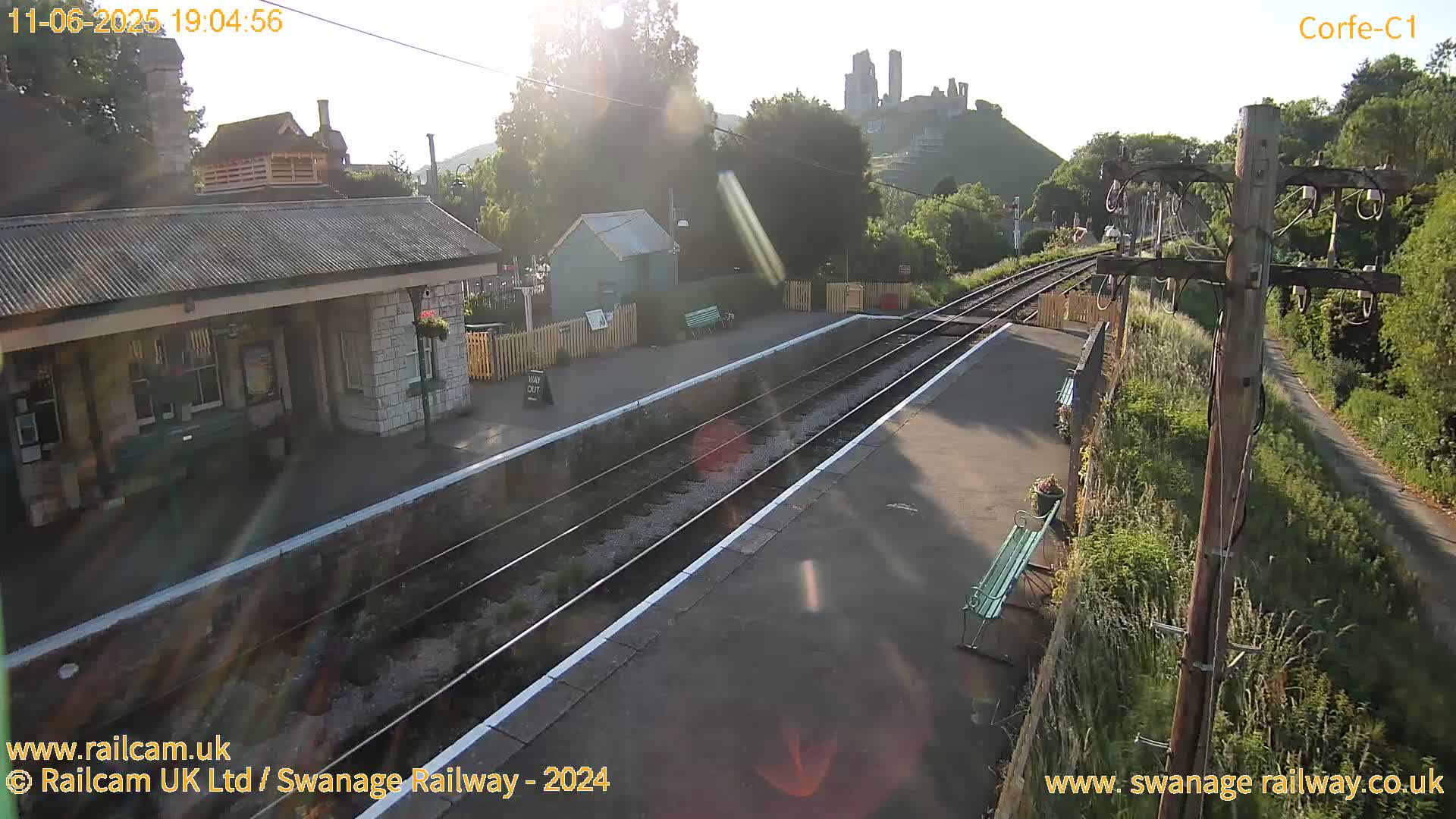 A train station platform with a single track and a stone building, situated beneath a hill with a ruined castle, on a sunny day.