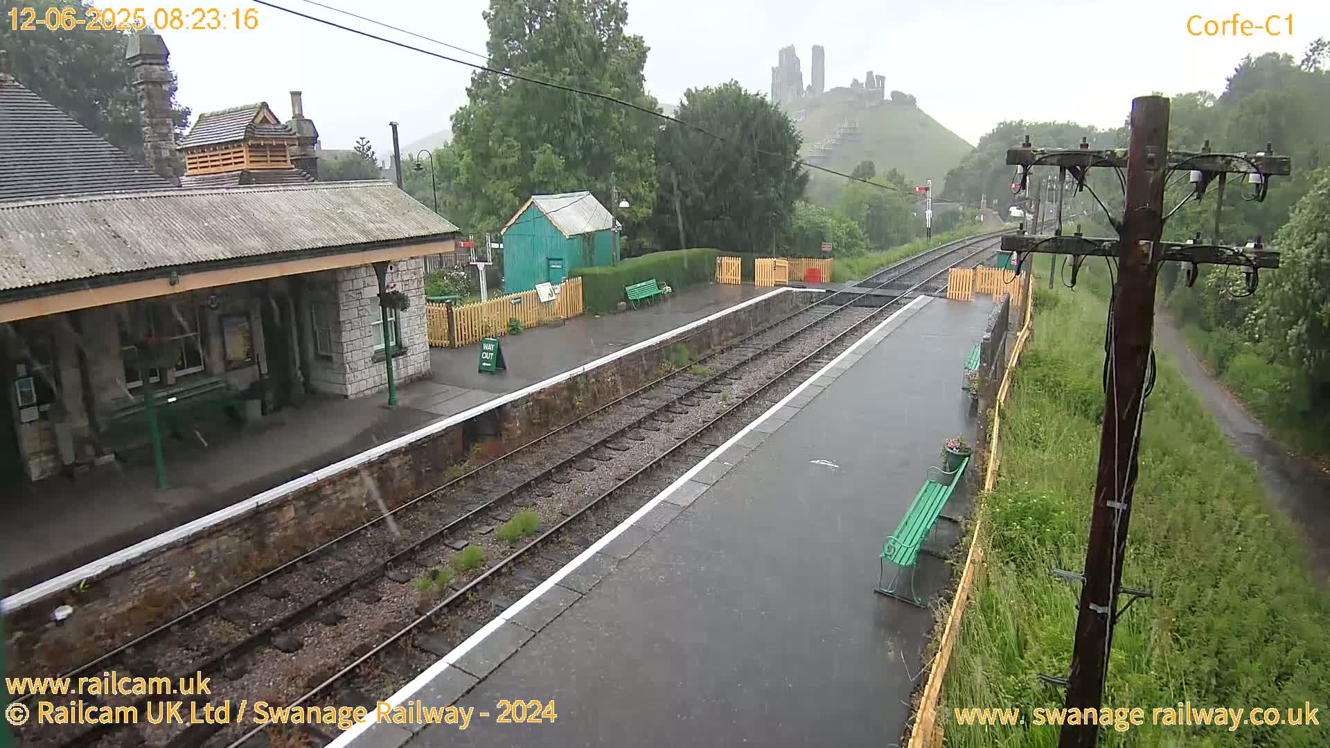 A rain-soaked railway station platform with a stone building, a small green shed, and a distant castle visible through the downpour.