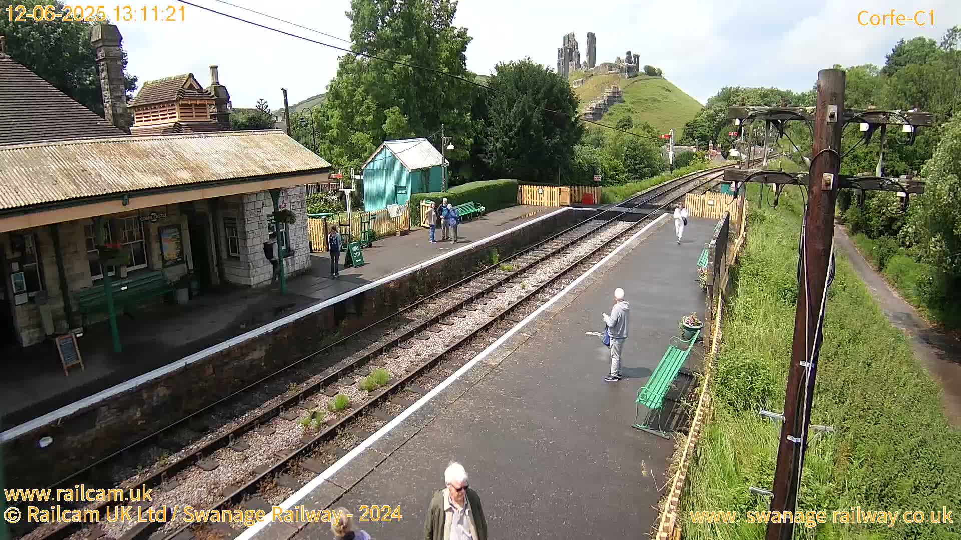A sunny day at a train station with a stone building, a small green shed, and a ruined castle visible in the distance.