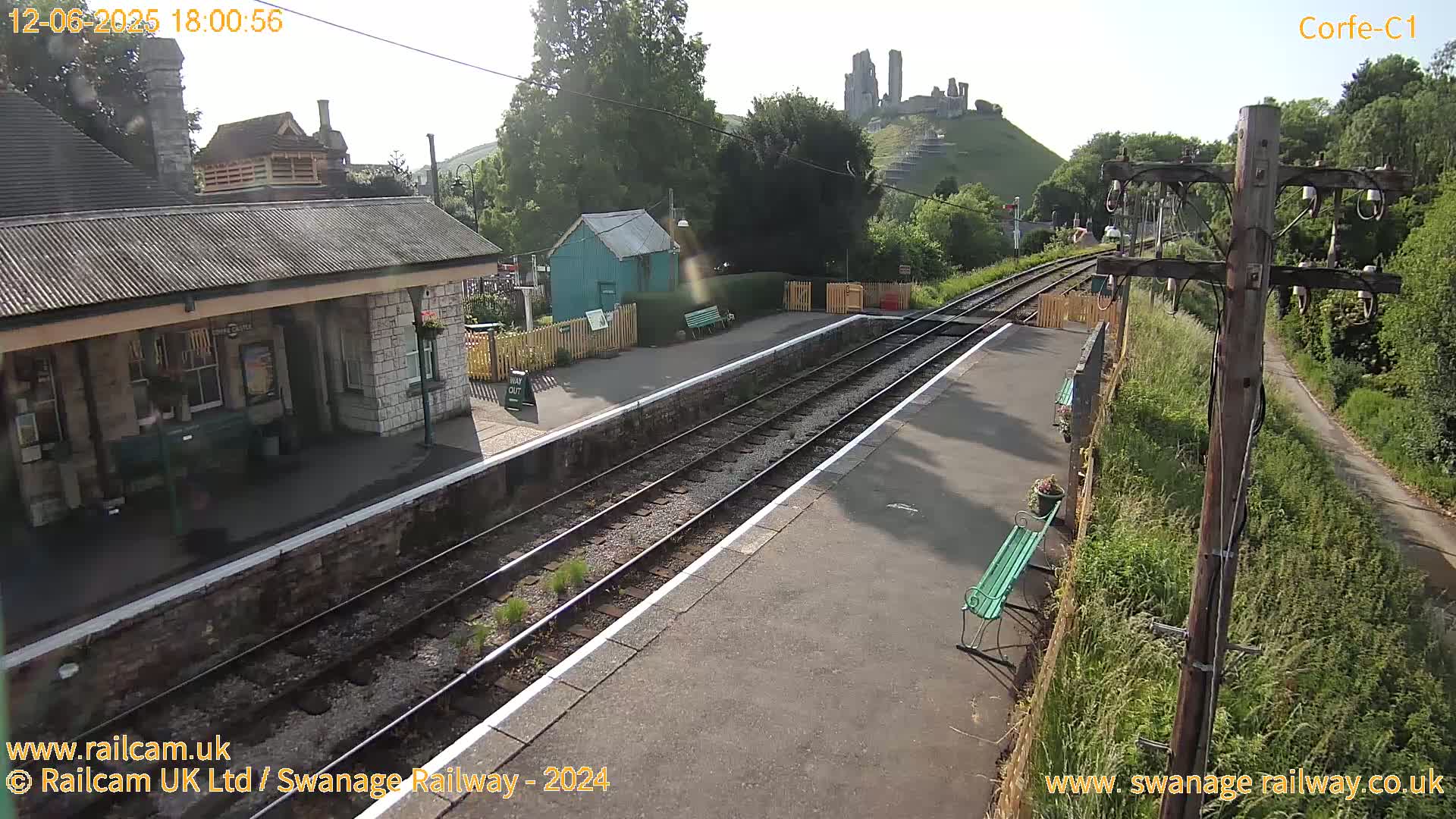 A sunny view of a train station platform with a stone building, a small teal shed, and a distant castle ruin on a hill.